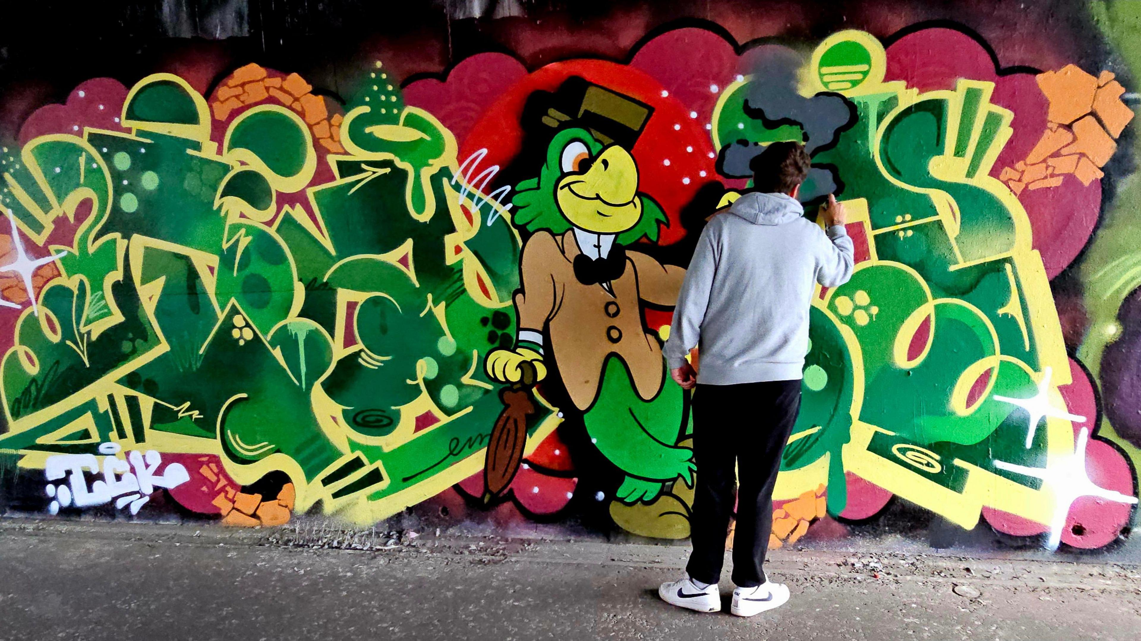 An artist working on a mural featuring a green parrot in a top hat with an umbrella