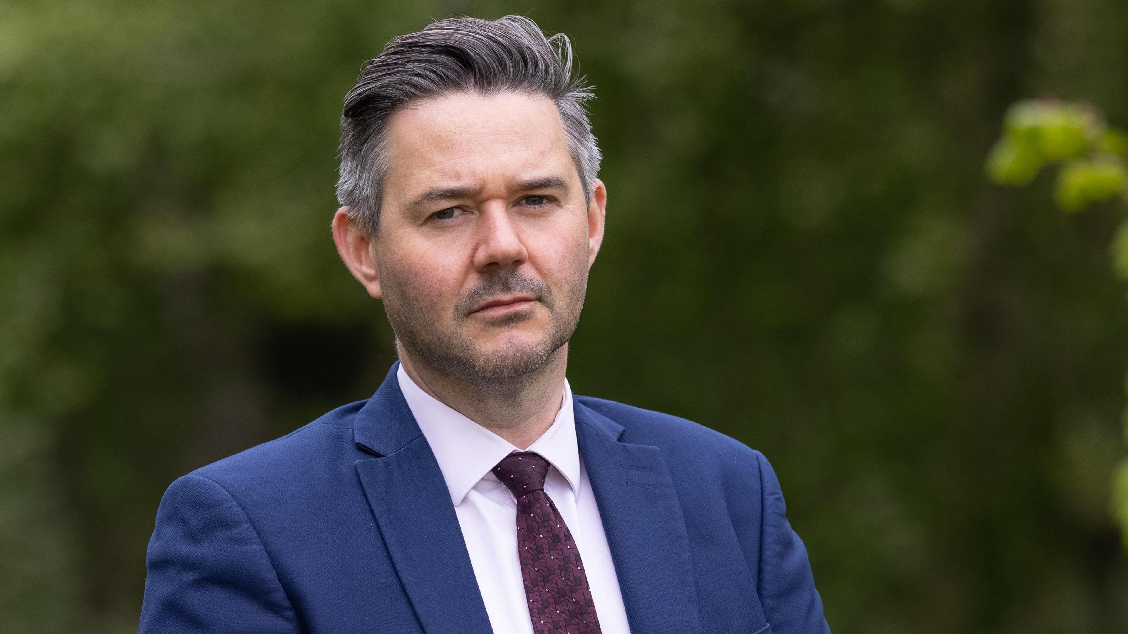 An unsmiling Oliver Burwood looks directly at the camera wearing a blue suit and burgundy tie.