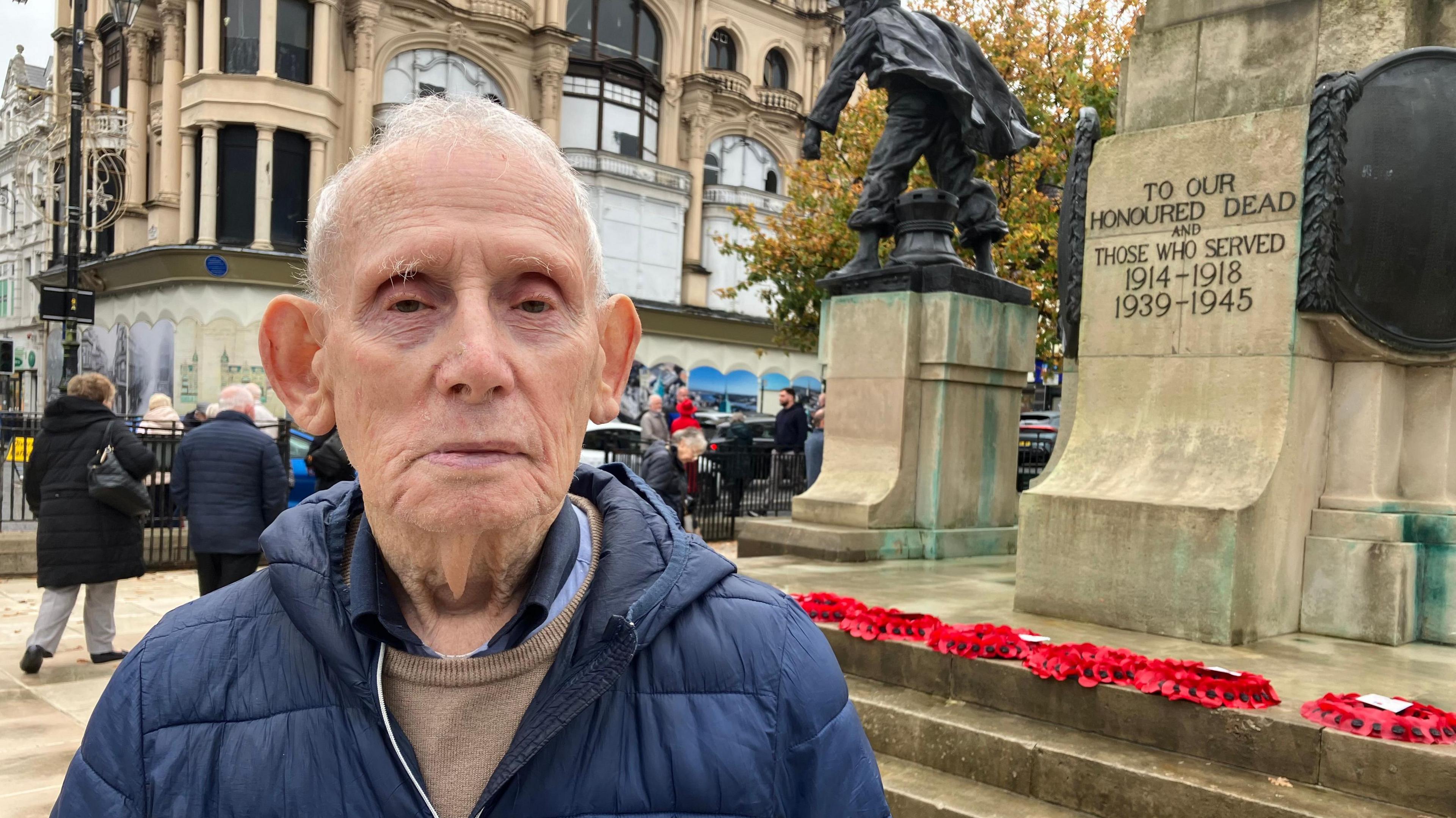 An elderly man wearing a shirt, jumper and navy coat looks at the camera. A cenotaph with poppy wreaths can be seen behind him as can other people in the background.  