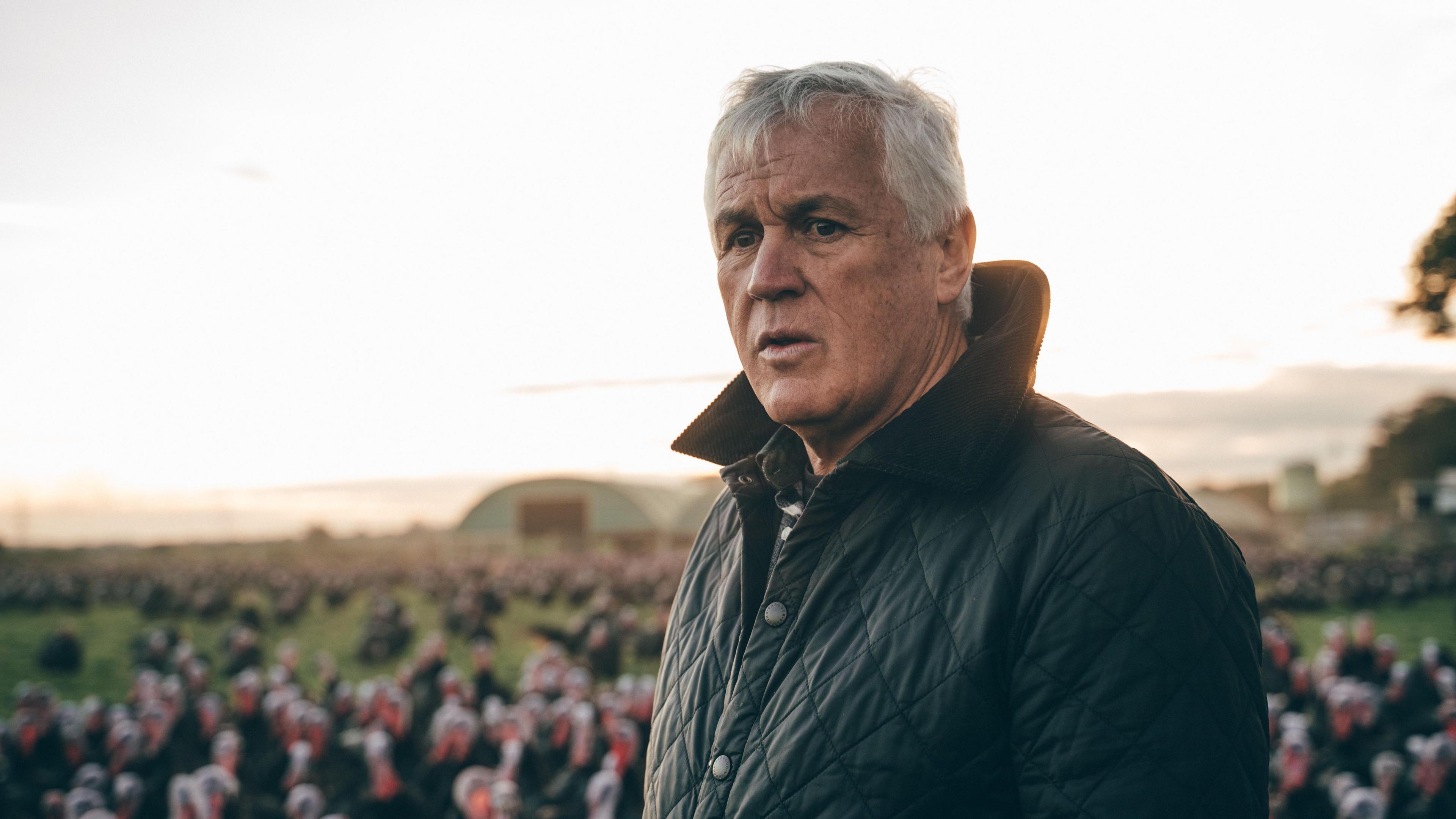 A farmer with medium length white hair and a dark green quilted jacket stands in a portait. In the background but blurred out of focus are dozens of turkeys. They are among green grass and there's a type of domed green outhouse in the far distance.