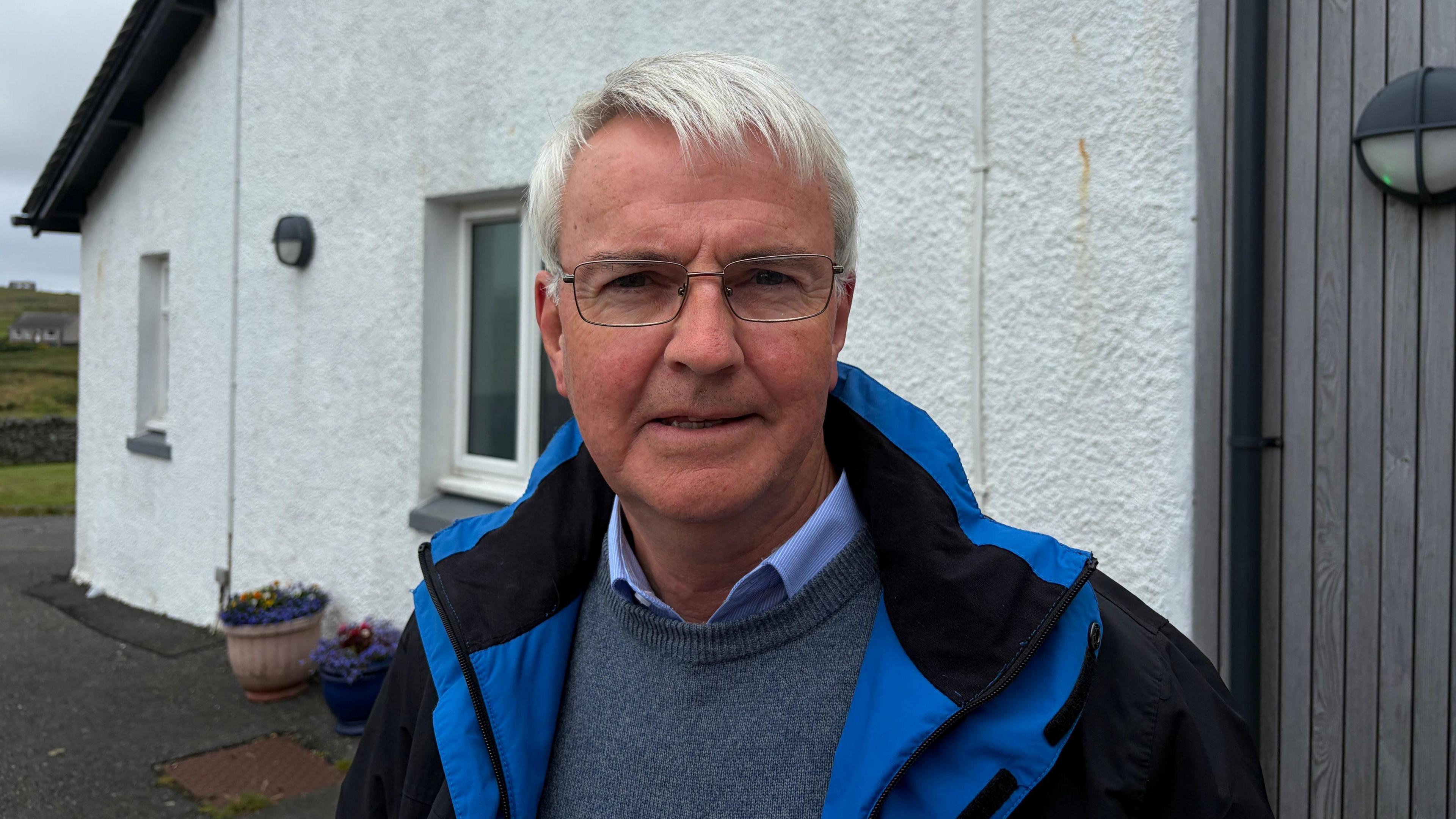 Aonghas Dòmhnallach. Fireannach le falt liath, speuclairean, seacaid, lèine agus geansaidh gorm
