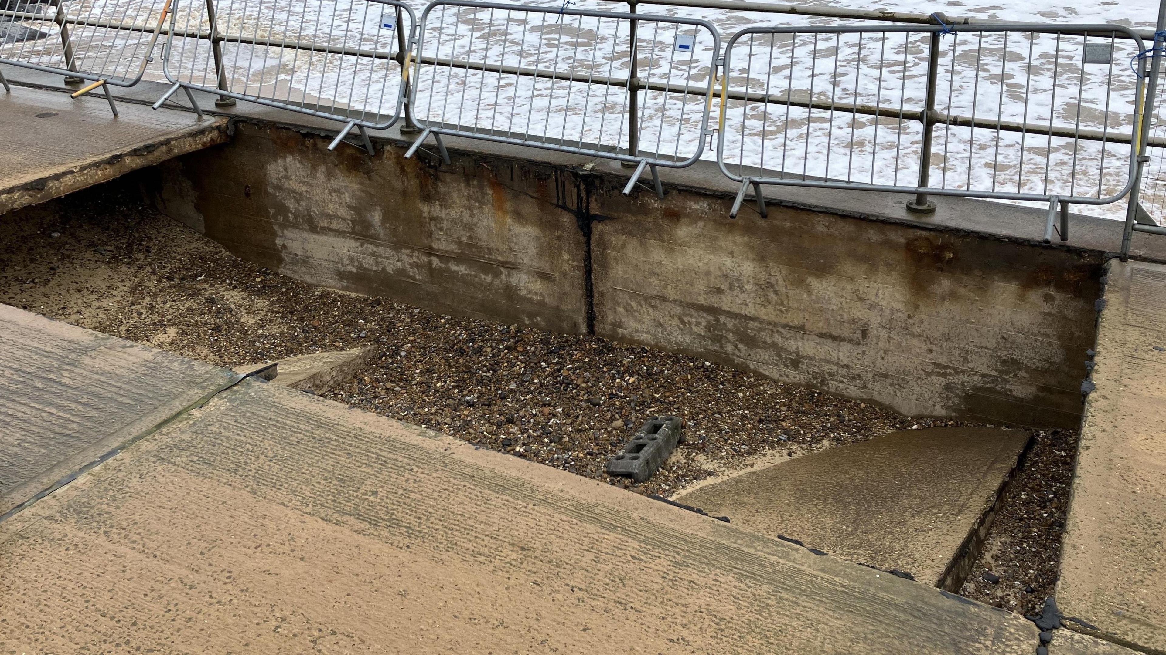 Metal fencing is in background and a large, rectangular-shaped hole in a concrete promenade has shingle inside and the remnants of the collapsed walkway.