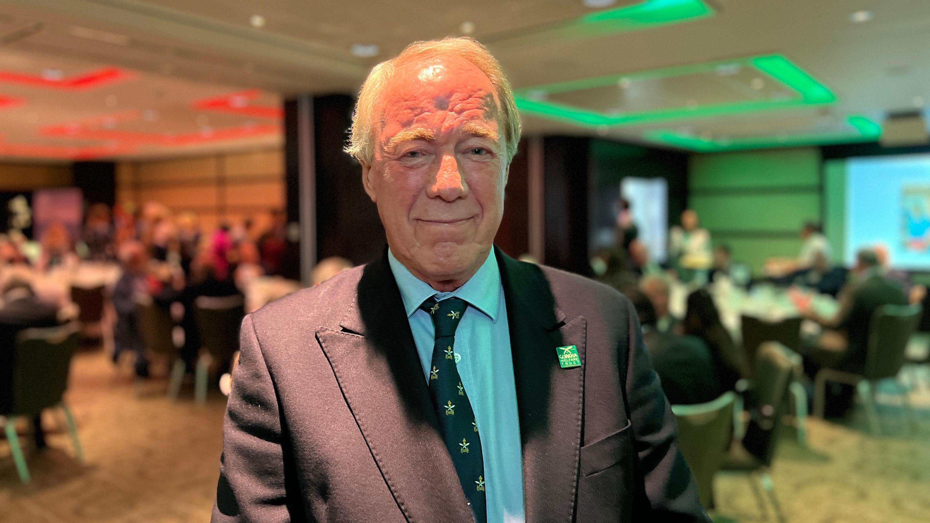 John looks at the camera as people are sitting down for dinner and getting food behind him. He's wearing a navy blue jacket with a white shirt along with a green tie with Gurkha Trust logos on. He also has a Trust badge on his jacket.