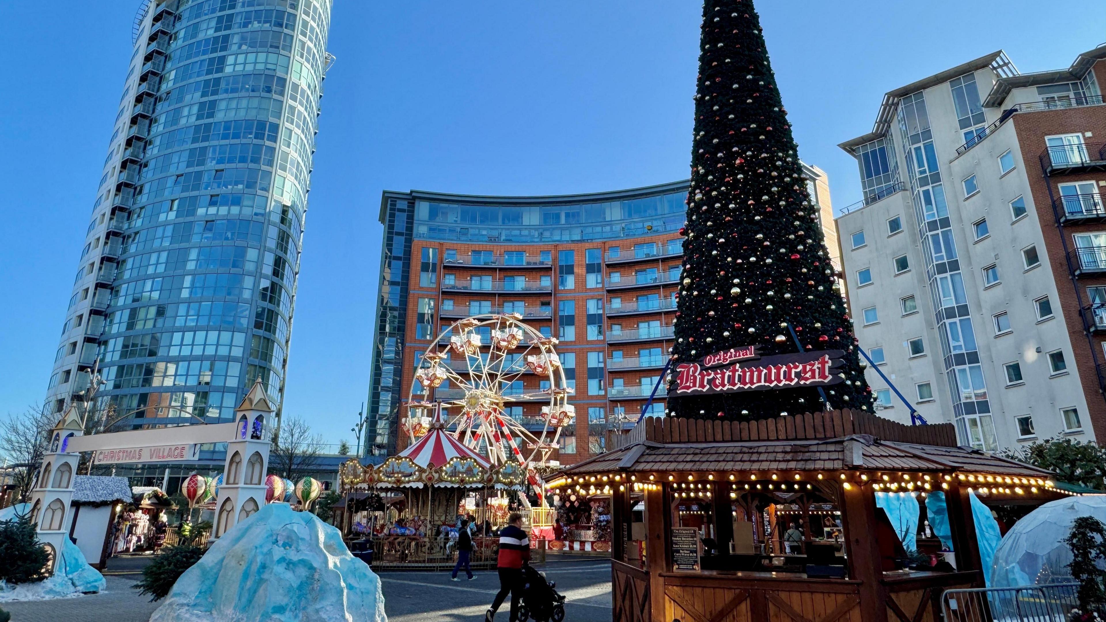 A city scene with a skyscraper and office block-looking buildings in the background. A Christmas market can be seen in the foreground with a small ferris wheel, Bratwurst stand and other stalls. Large plastic icebergs are also scattered on the ground in the market.