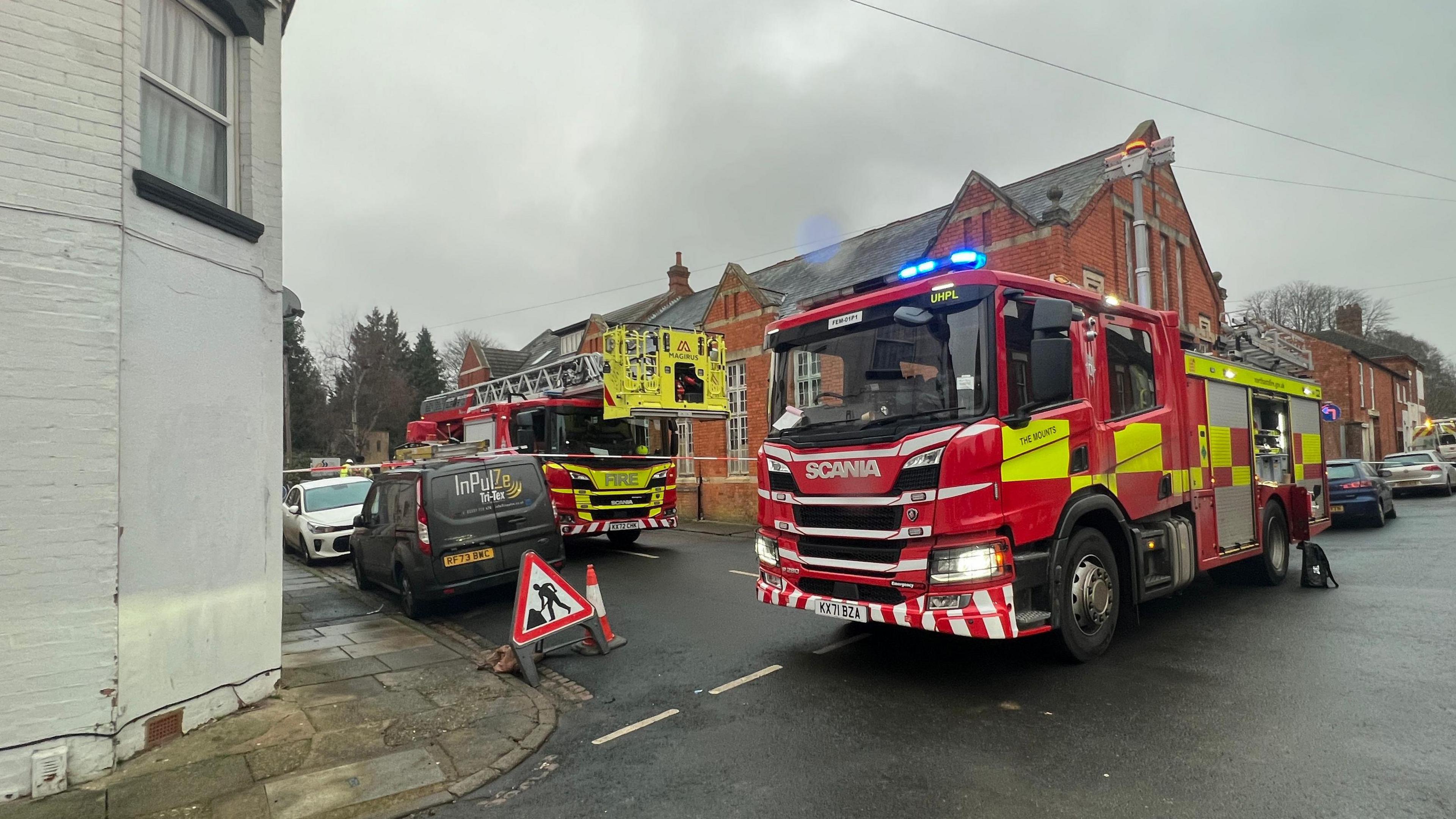 Two fire engines are parked alongside and in front of a red brick building in the middle of the intersecting roads. The roads are wet and the sky grey.