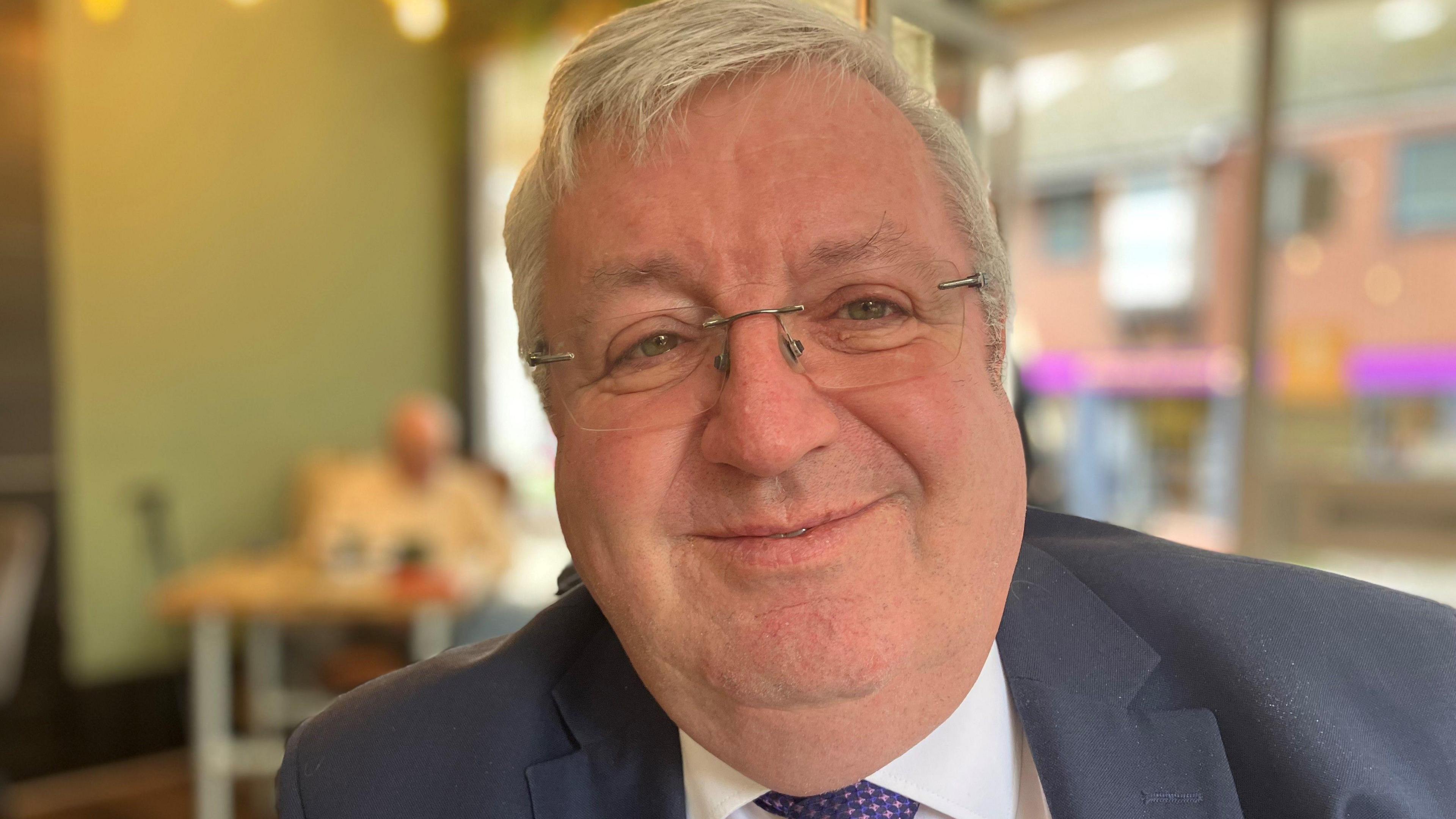 A man with short white hair, glasses and wearing a suit, smiles while he is sitting in a cafe.