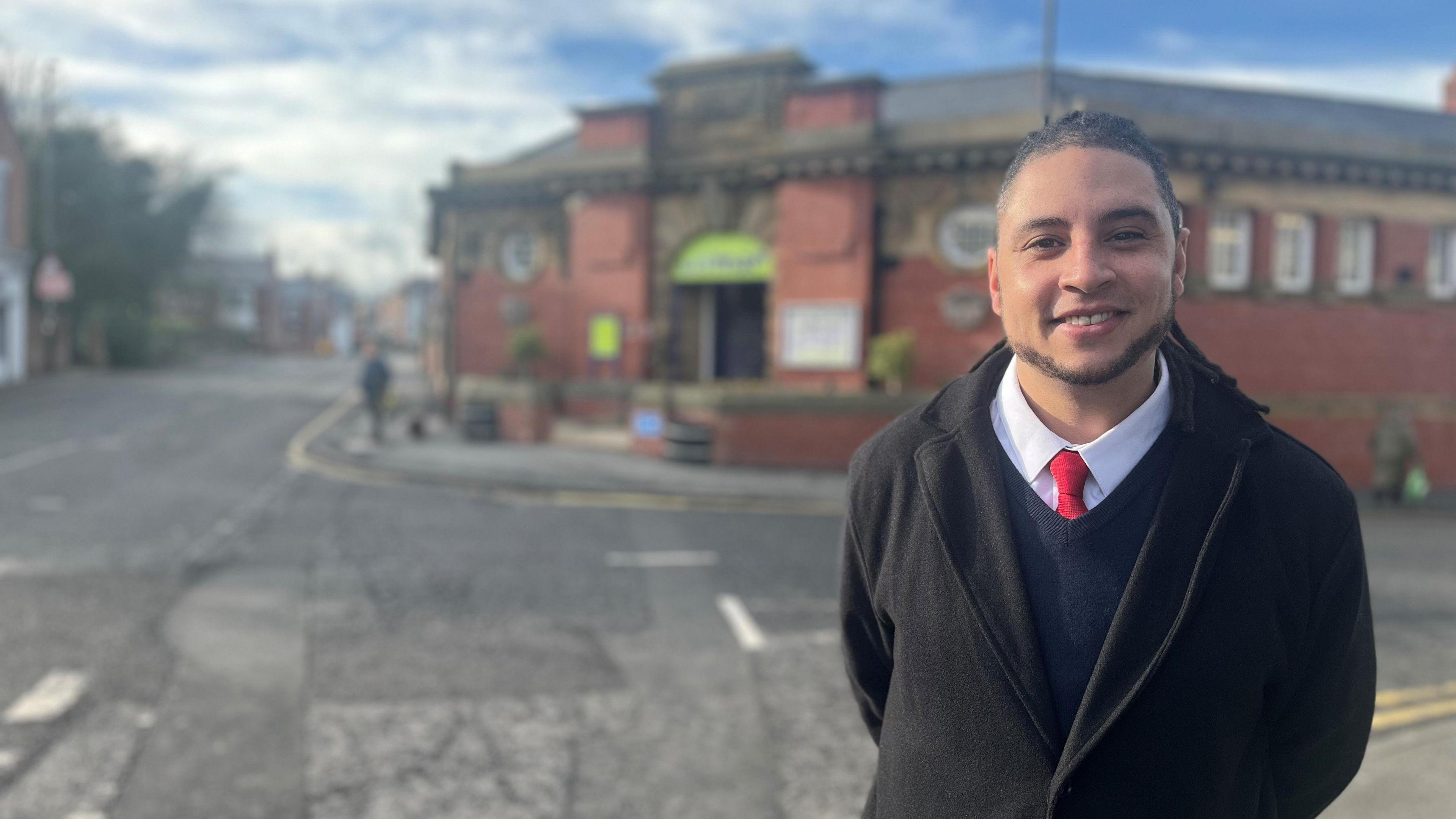 Stephen is smiling at the camera. His braided hair is put back in a ponytail. He is wearing a black coat and navy blue jumper over a white shirt and red tie. 