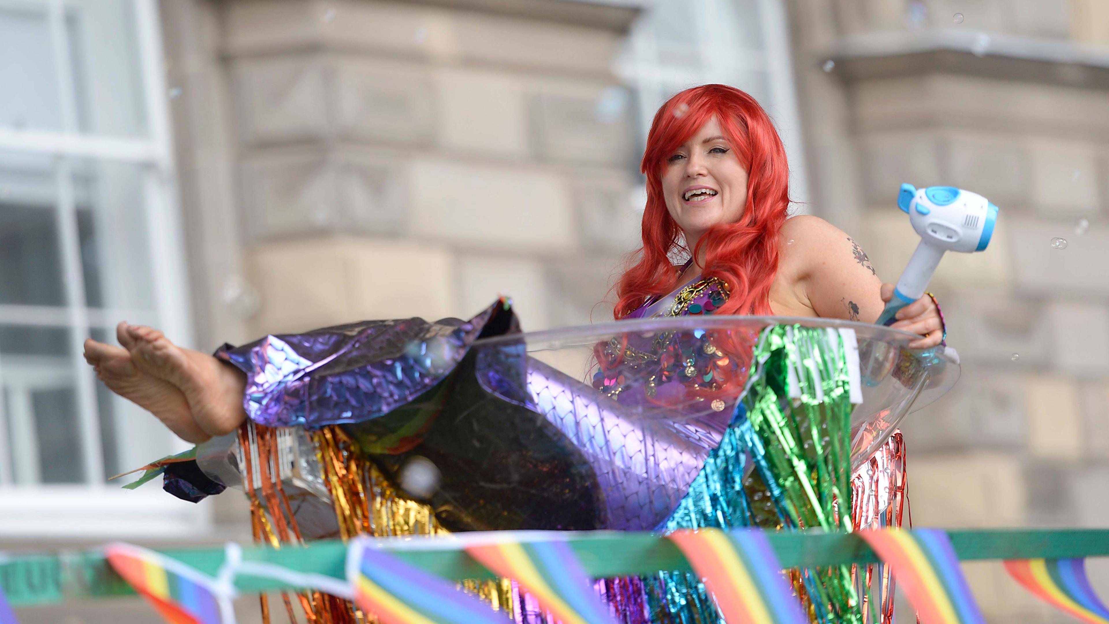 A woman dressed up as a mermaid is sitting in a large plastic bowl. She is wearing a long red wig and has a purple shiny costume which looks like a mermaid's tail.