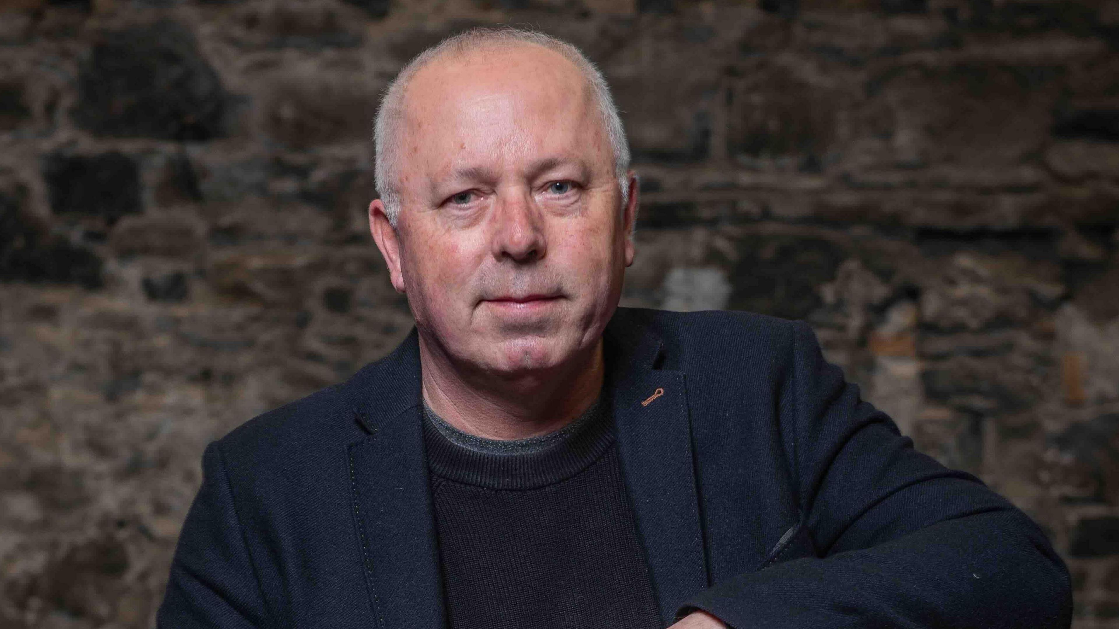 A bald man in a black blazer and jumper sitting down against a brick wall. He has blue eyes and is half smiling at the camera.