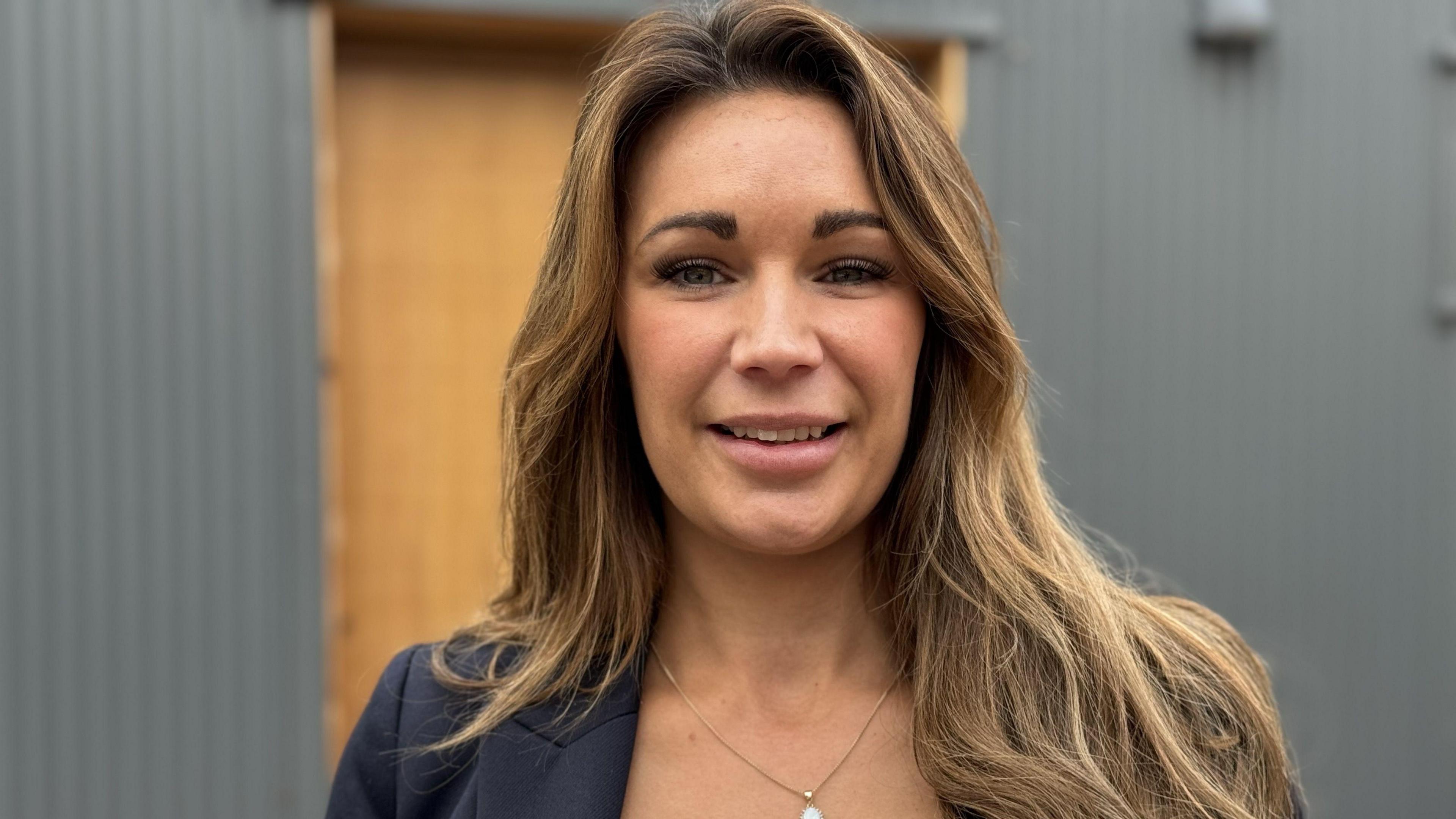 Gemma Parsons, a woman with brown hair, is standing outdoors in front of a grey building with a wooden door. She is smiling while facing directly into the camera.