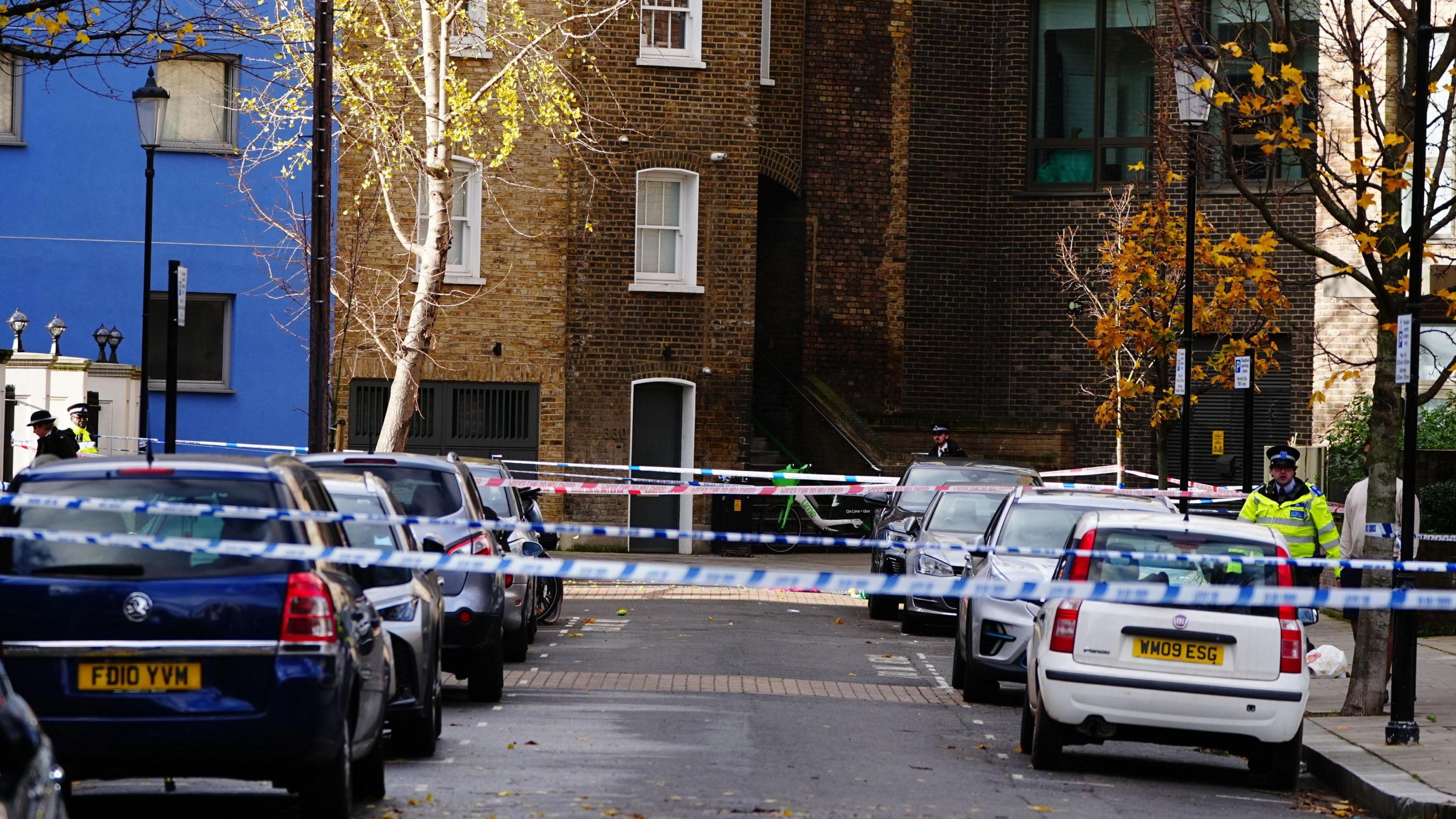 A police cordon at the scene on Southern Grove in Ladbroke Grove. A row of blue, silver and white cars can be seen on either side of the street which has five blue, red and white police cordons criss-crossing the road