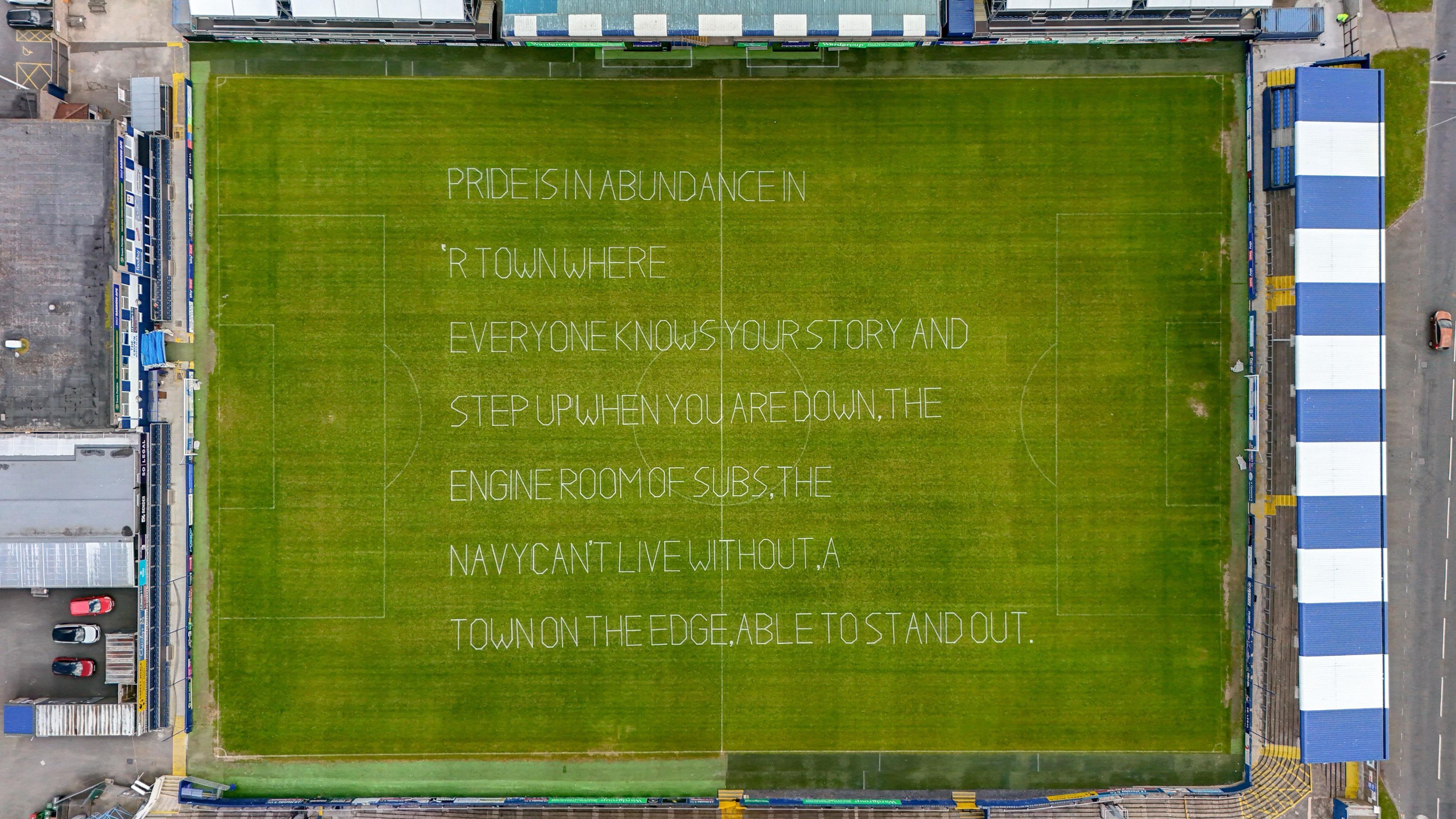 Shot from Colin Aldred, accredited drone user: Aerial view directly above the pitch at Holker Street, showing a poem in white lettering on the grass