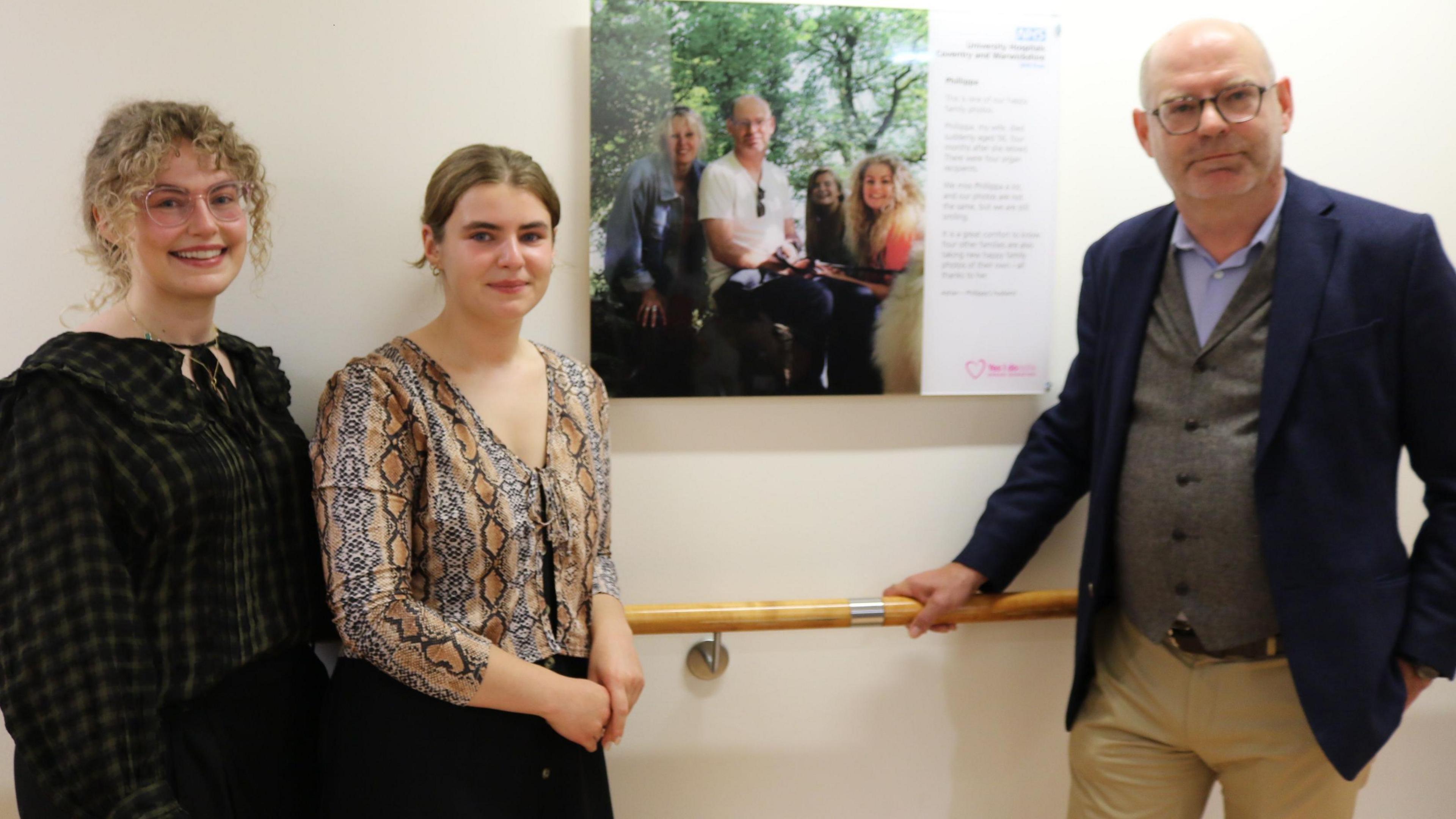 Two women standing to the left of a wall-mounted poster in a corridor, with a man on the right. One woman is wearing a checked blouse and the other a snakeskin printed one, while the man is wearing a three-piece suit.