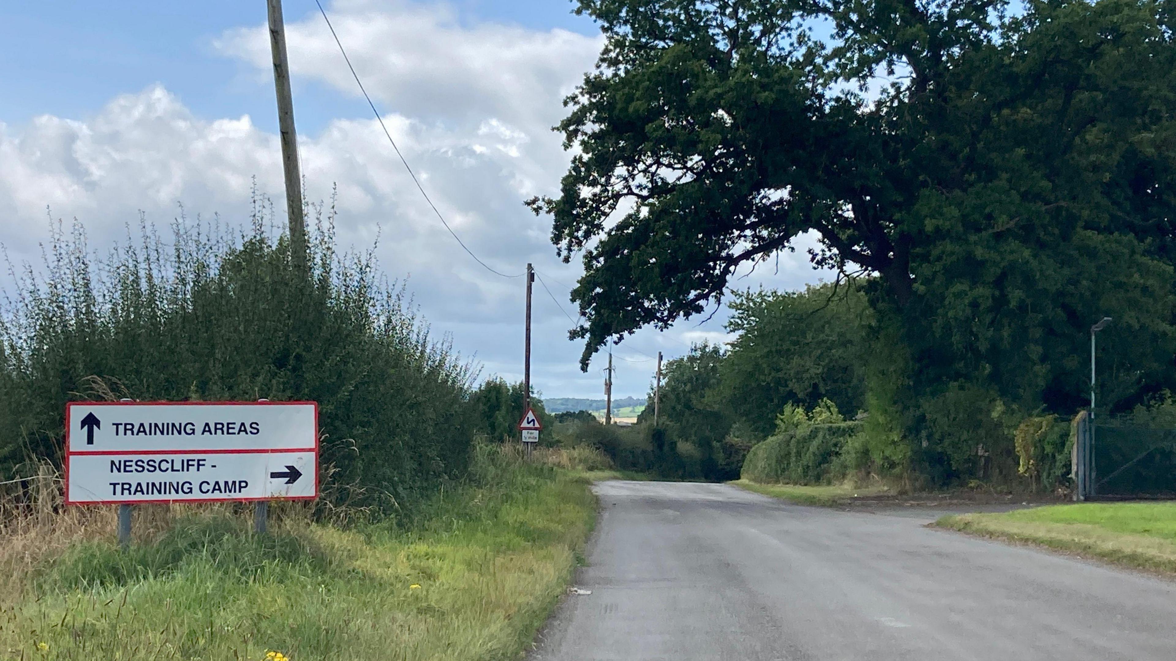 A country road, surrounded by green hedges and trees. There is a sign on the left that says "Nescliffe training camp" with an arrow pointing to the right.