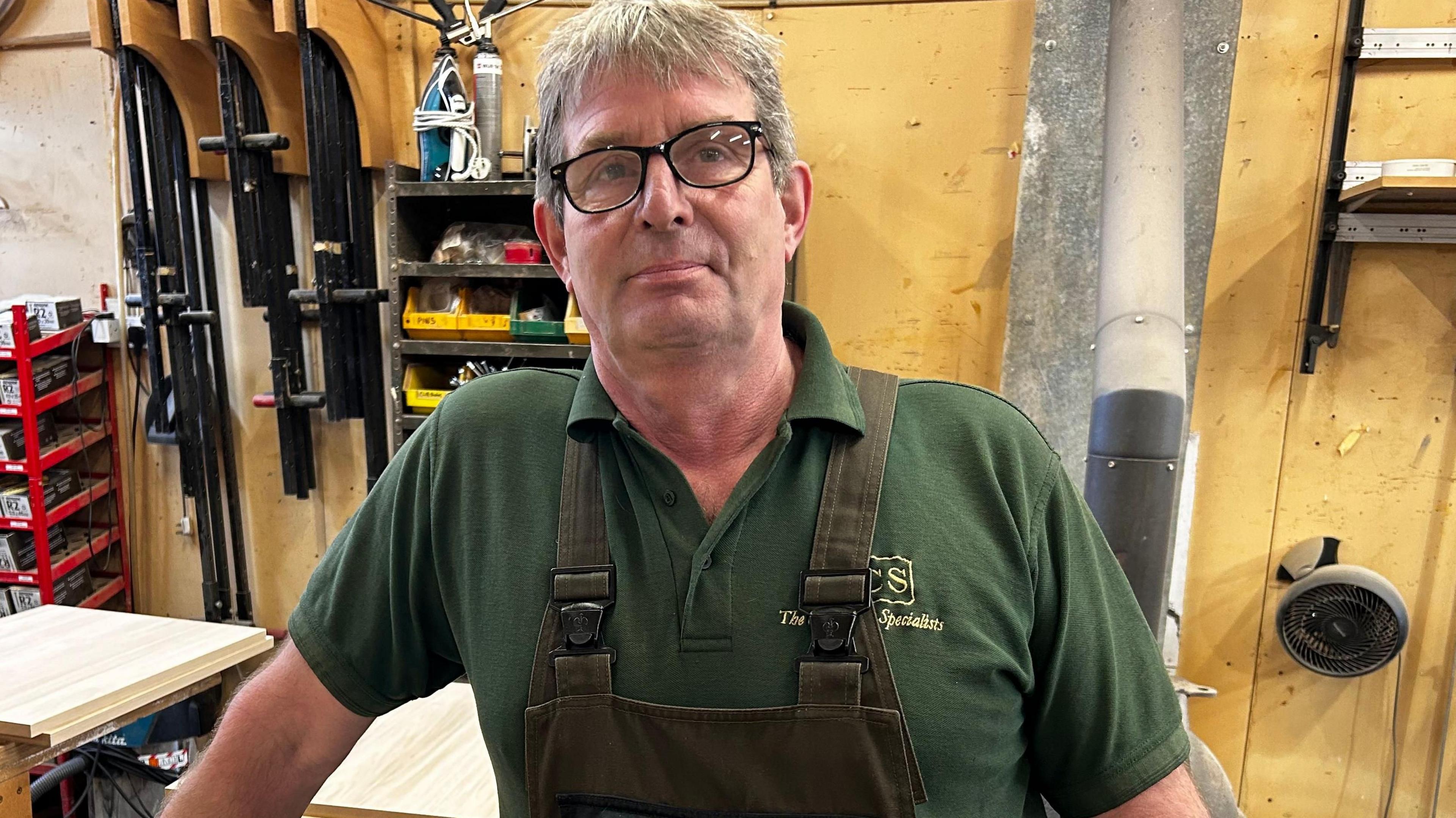 Steve Davies is seen in a green polo shirt and overalls leans on a workbench in a carpentry workshop, with wooden panels and tools behind him.