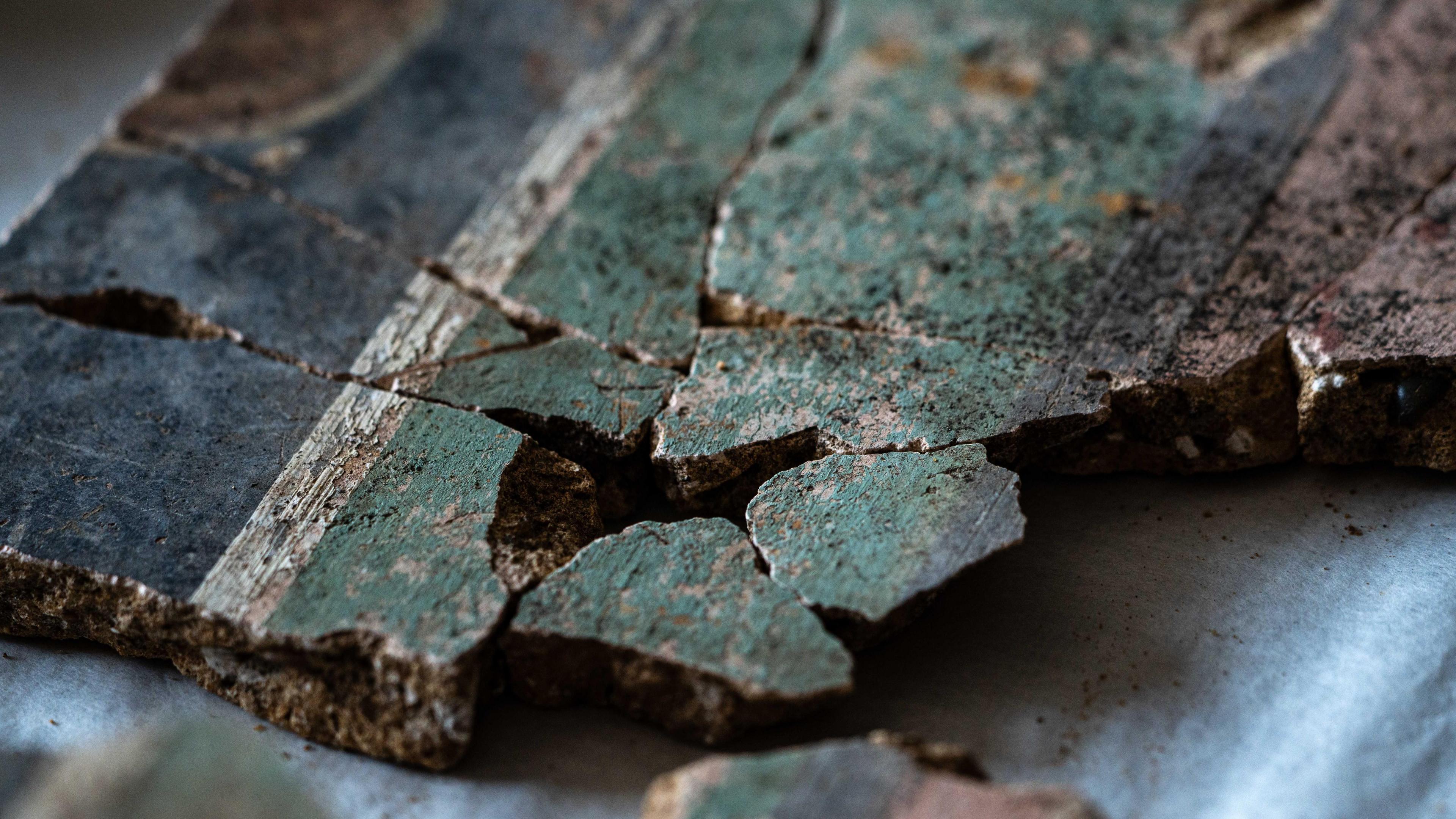 A close up image of at least 10 fragments of the plaster that have been pieced together. On the right hand side of the image is the pink dado section, then a thin black dividing line and a thicker pale green band. Towards the left of the image is a thin white dividing line and then a thicker black band.