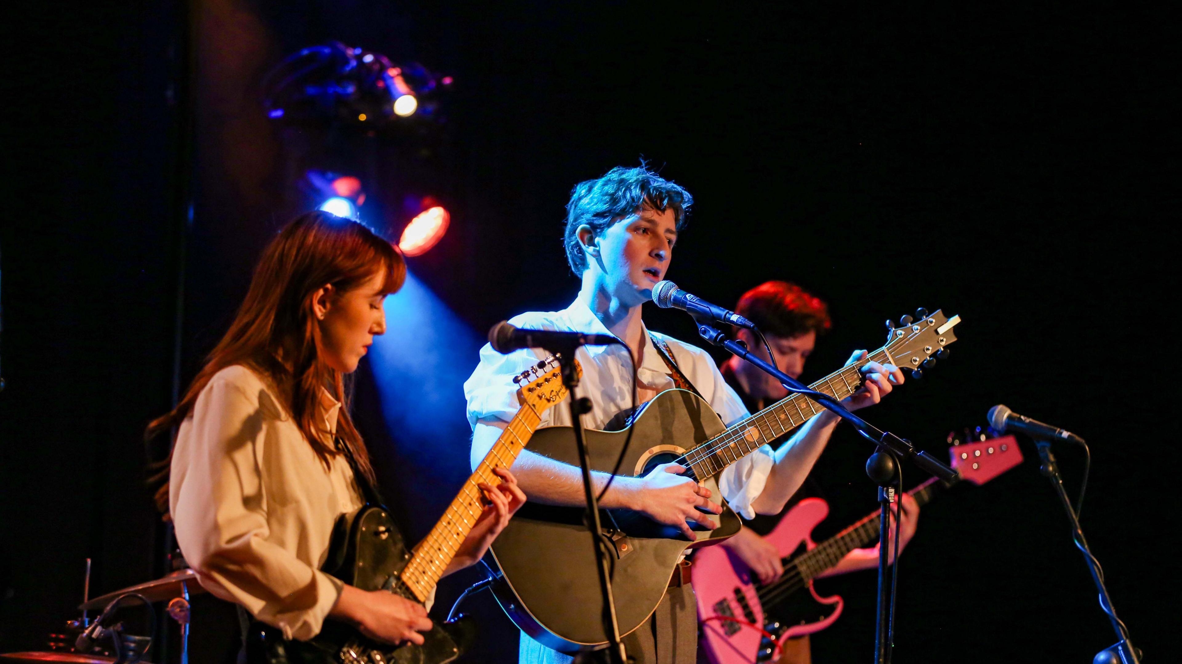 Two men and a woman are stood on stage performing. The woman is wearing a white shirt and has long brown hair. She is holding an electric guitar. The first of the two men is stood in the middle of the three. He is wearing a white shirt and is playing an acoustic guitar. He is also singing into the microphone. The second man is playing a pink guitar. 