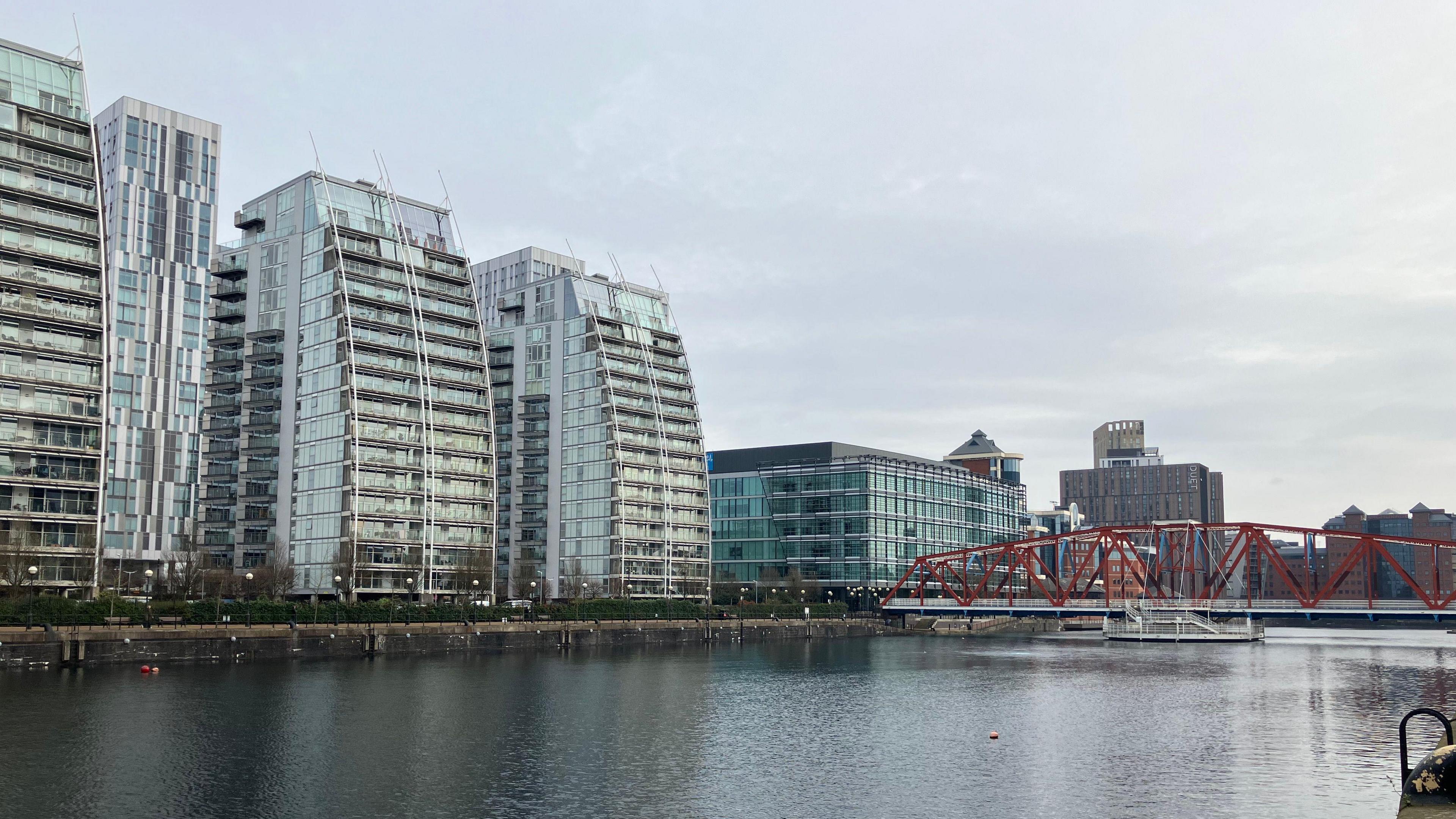 Tall, glass buildings are surrounding a body of water. There is a red bridge running over the water. 