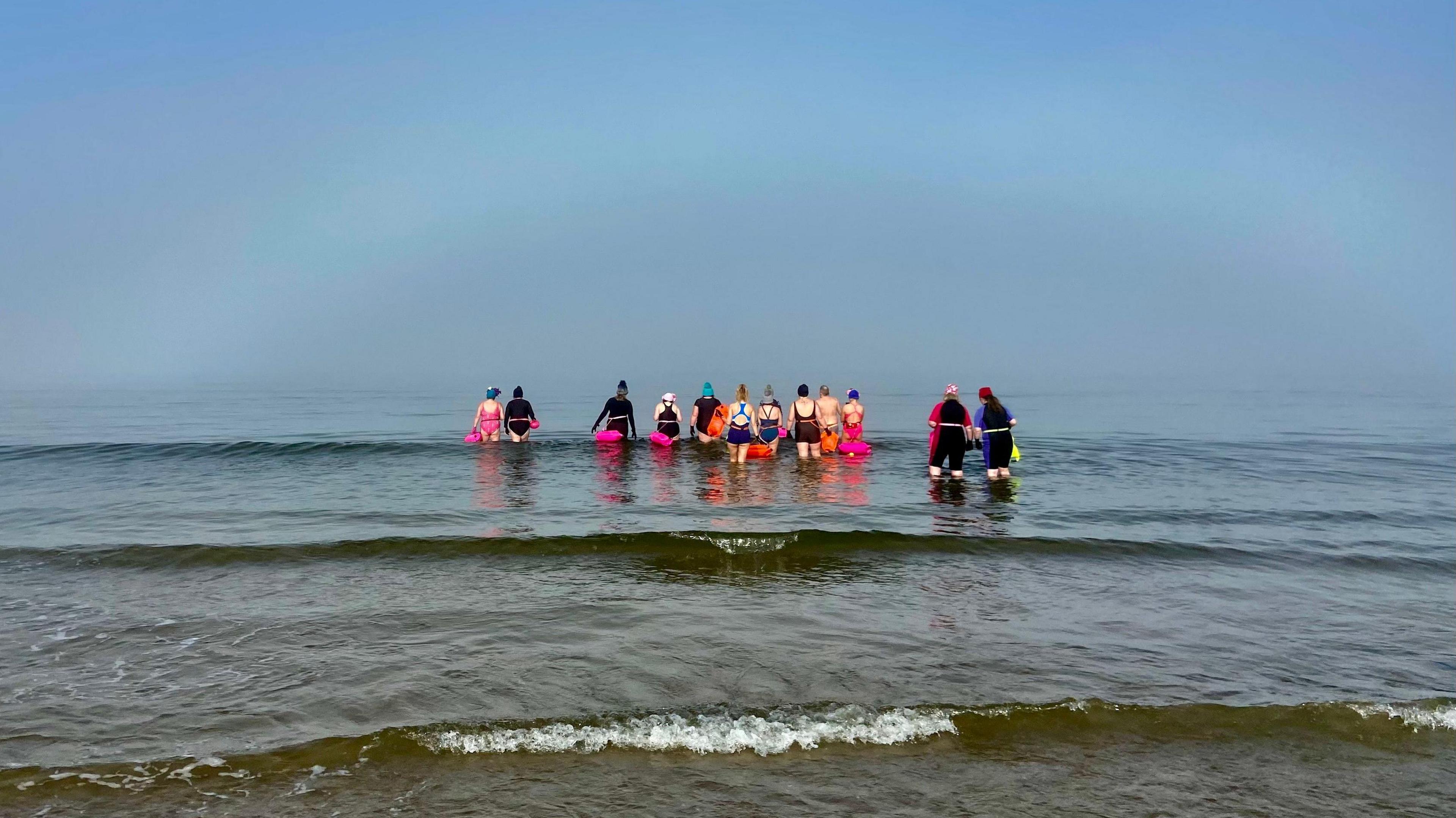 A group of 12 people in the water at Nairn Beach. Most are wearing swimsuits in various colours but four are wearing full body wetsuits in dark colours. All are wearing swimming caps. The water is calm and blue coloured, like the sky. A wave with white foam is rolling in in the foreground.