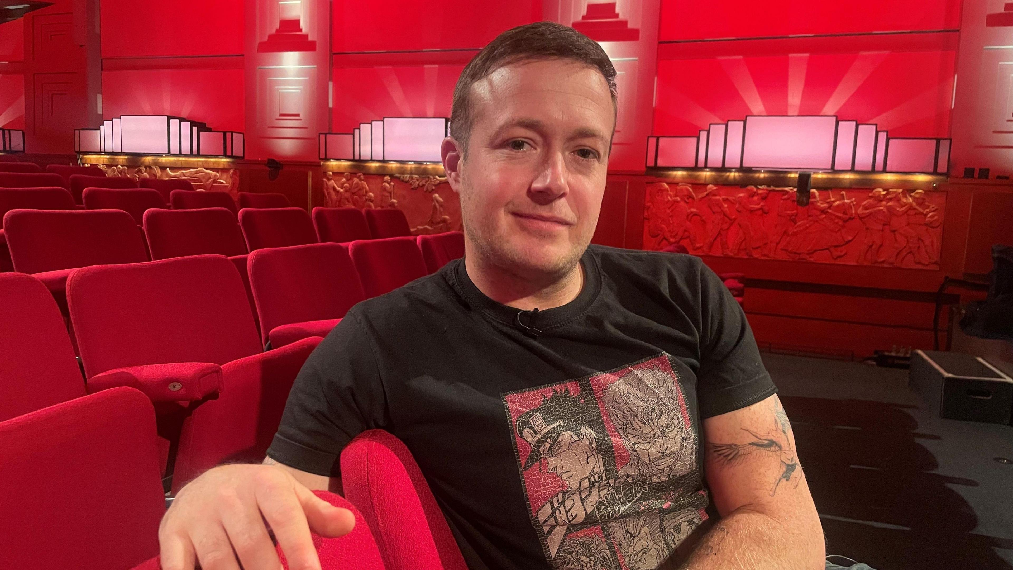 Jack Bence, wearing a T-shirt featuring anime figures, is pictured in the Radio Theatre in New Broadcasting House, sat on red theatre seats with Art Deco-style lighting lining the red wall behind him