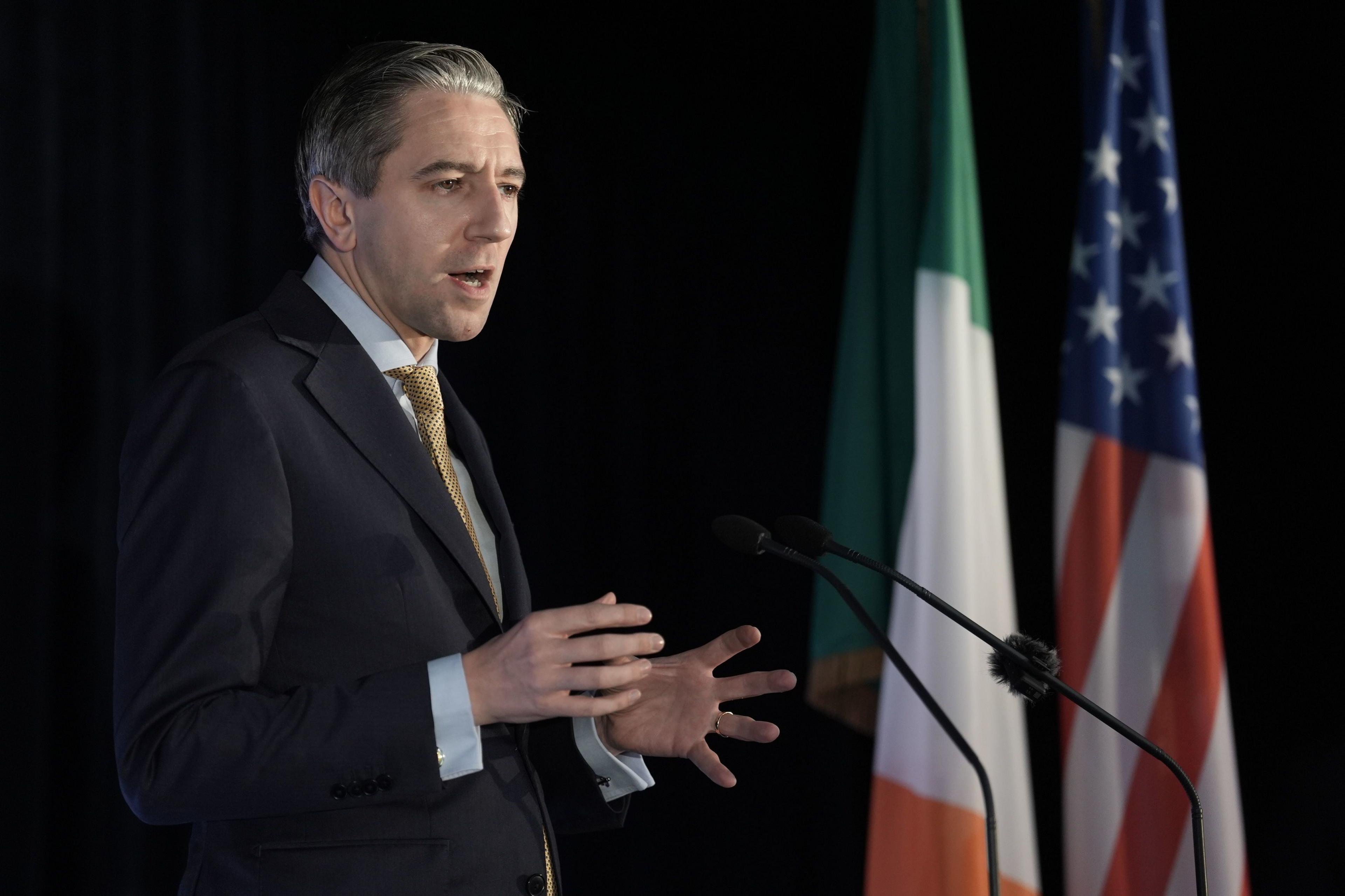 Simon Harris in grey suit jacket, blue shirt and gold tie, stood in front of a USA and Ireland flags, speaking into microphones at press event