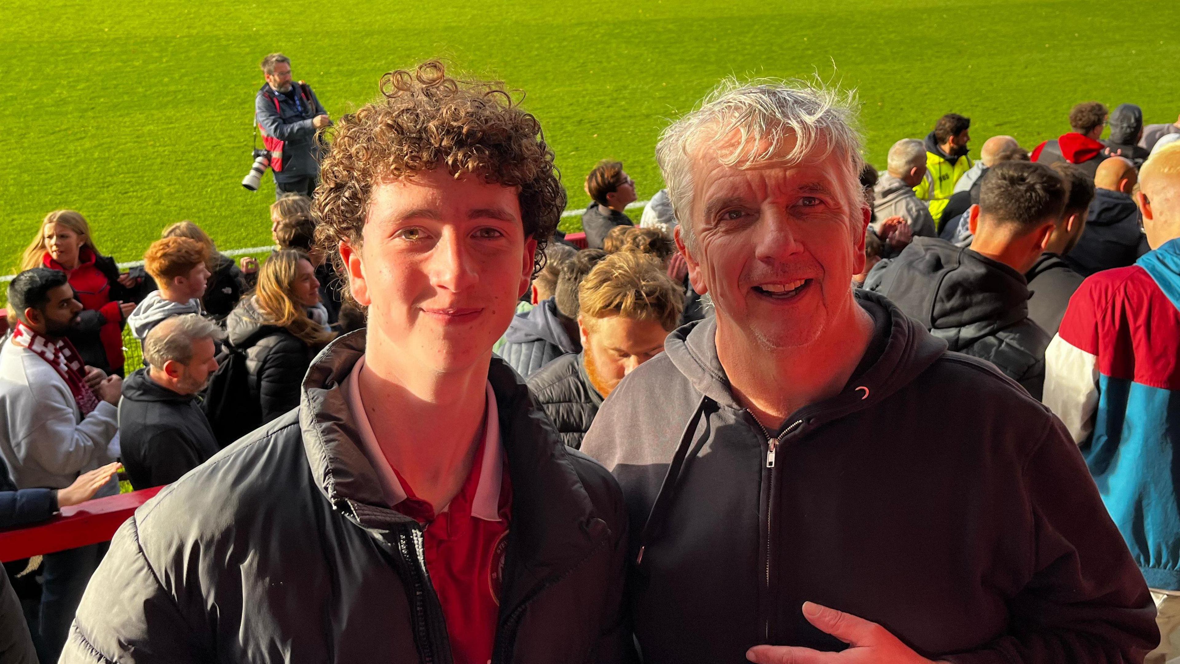 Oscar and John at a Stevenage FC game, with a large crowd of people behind them. They are both smiling and looking at the camera. A football pitch is behind them, with a camera man on the pitch. 