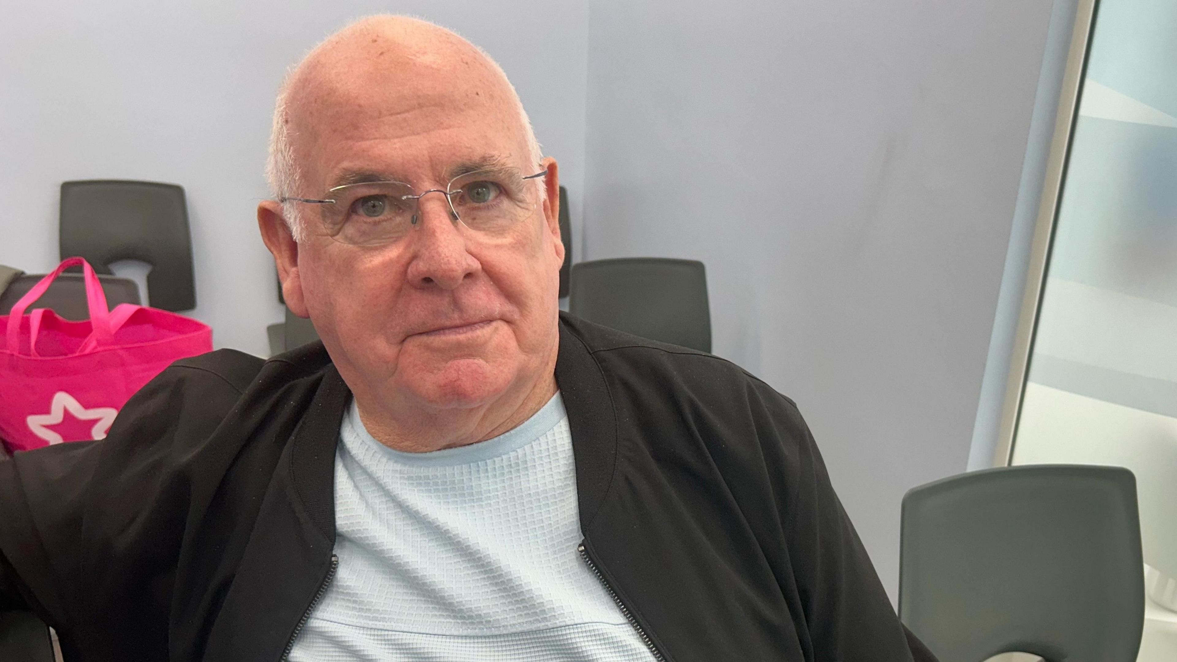 A man with a shaved head and glasses is looking at the camera. He is sitting amongst plastic chairs in a waiting room. There is a pink bag behind him.