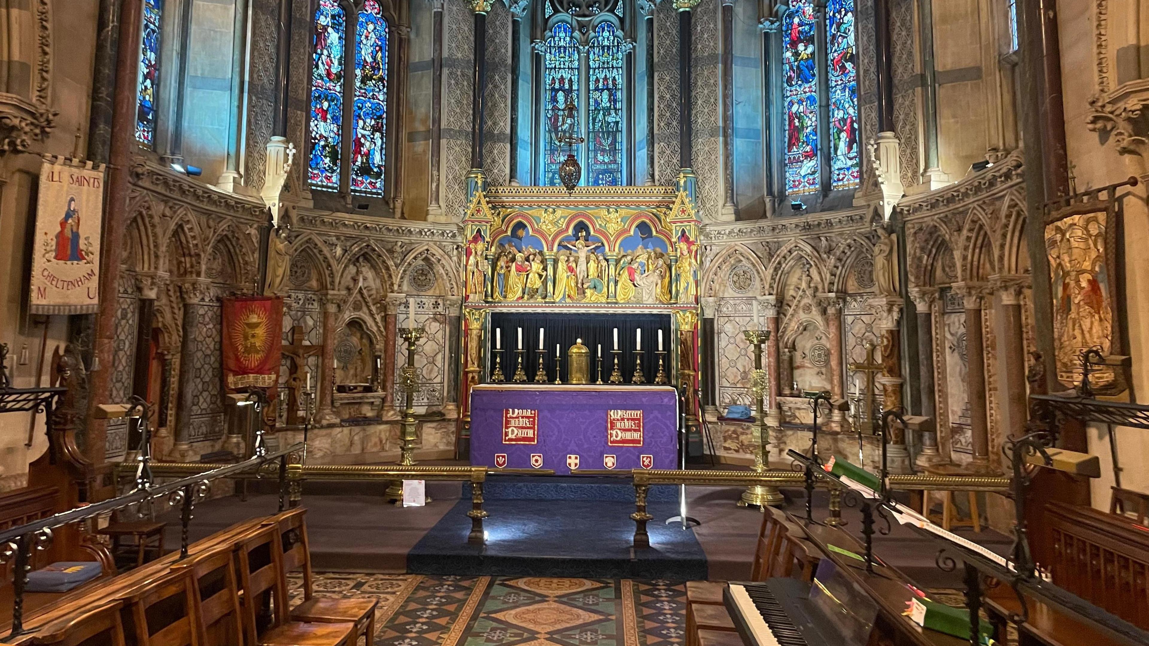 Image of the alter at the front of a church. The alter is dressed in purple and the walls are adorned with gold artwork.