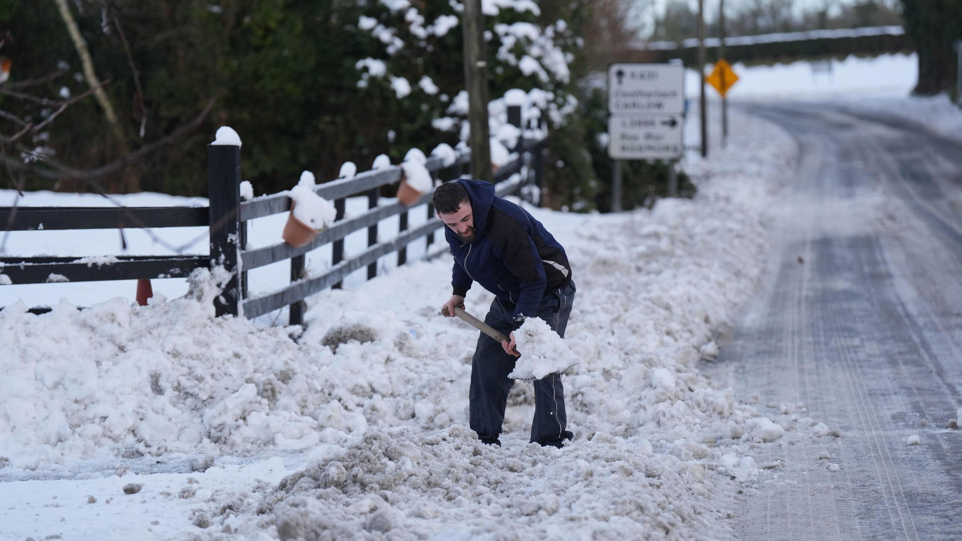 A man in a navy coat and navy tracksuit bottoms, shovels snow of the road. The man has black hair and a short beard. 