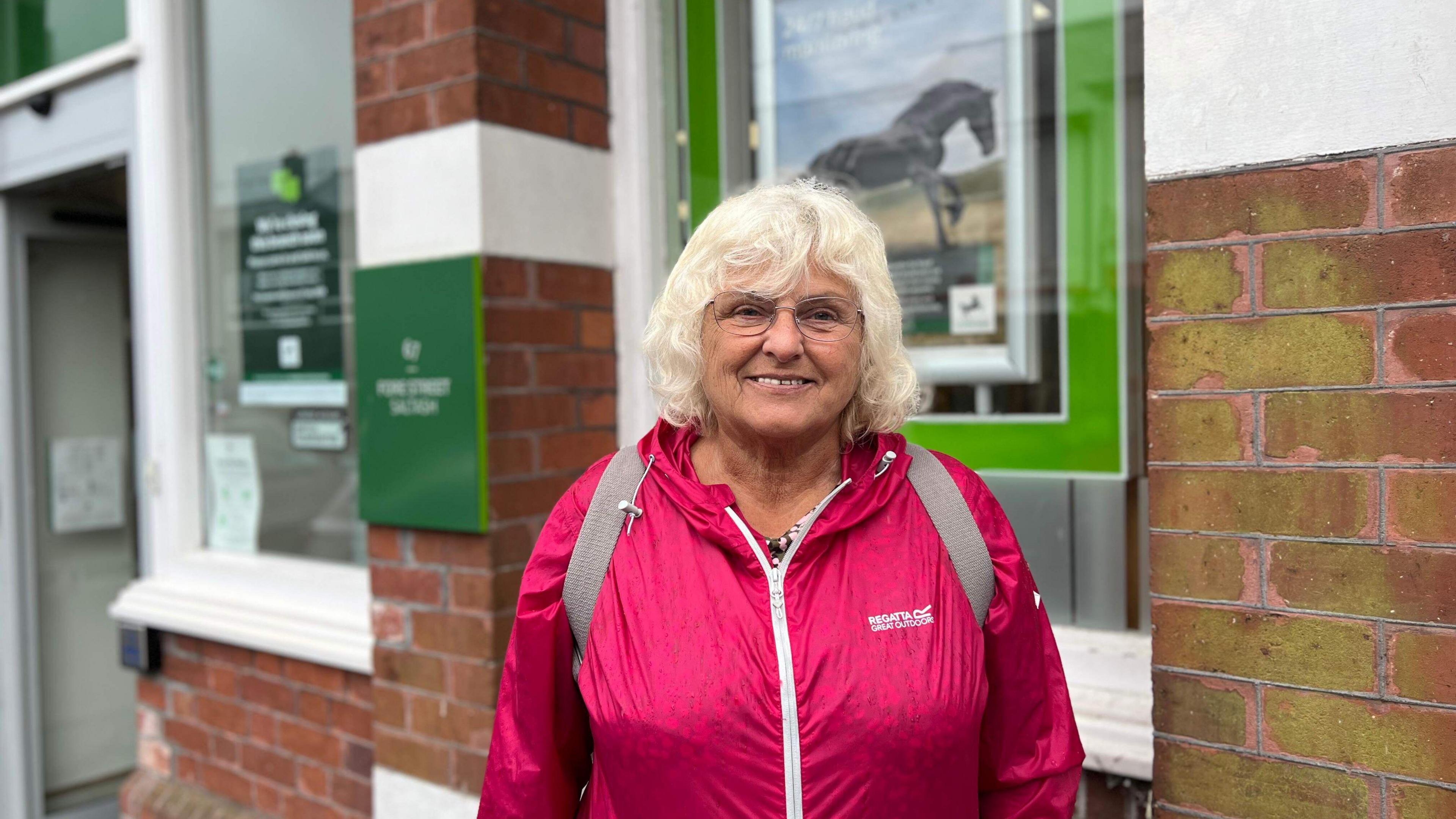 A woman with short blond hair, bright pink rain coat, glasses and rucksack looks straight at the camera smiling. Lloyds Bank signage can be seen slightly blurred in the background