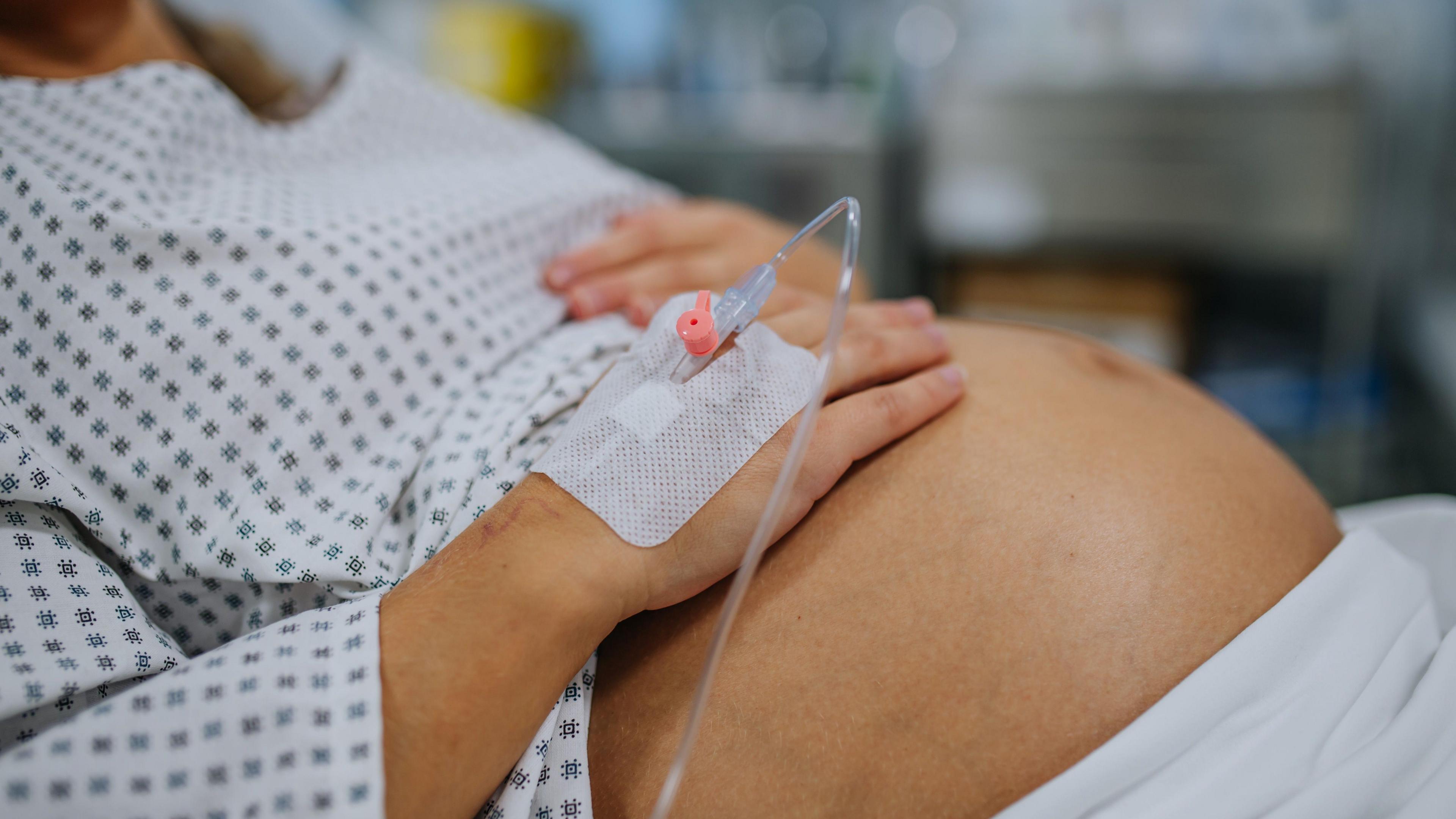 Close up of pregnant woman’s belly. Woman in labour waiting for examination in maternity ward. Woman during early stage of childbirth at the hospital holding her belly, feeling painful contractions. She's wearing a hospital gown.