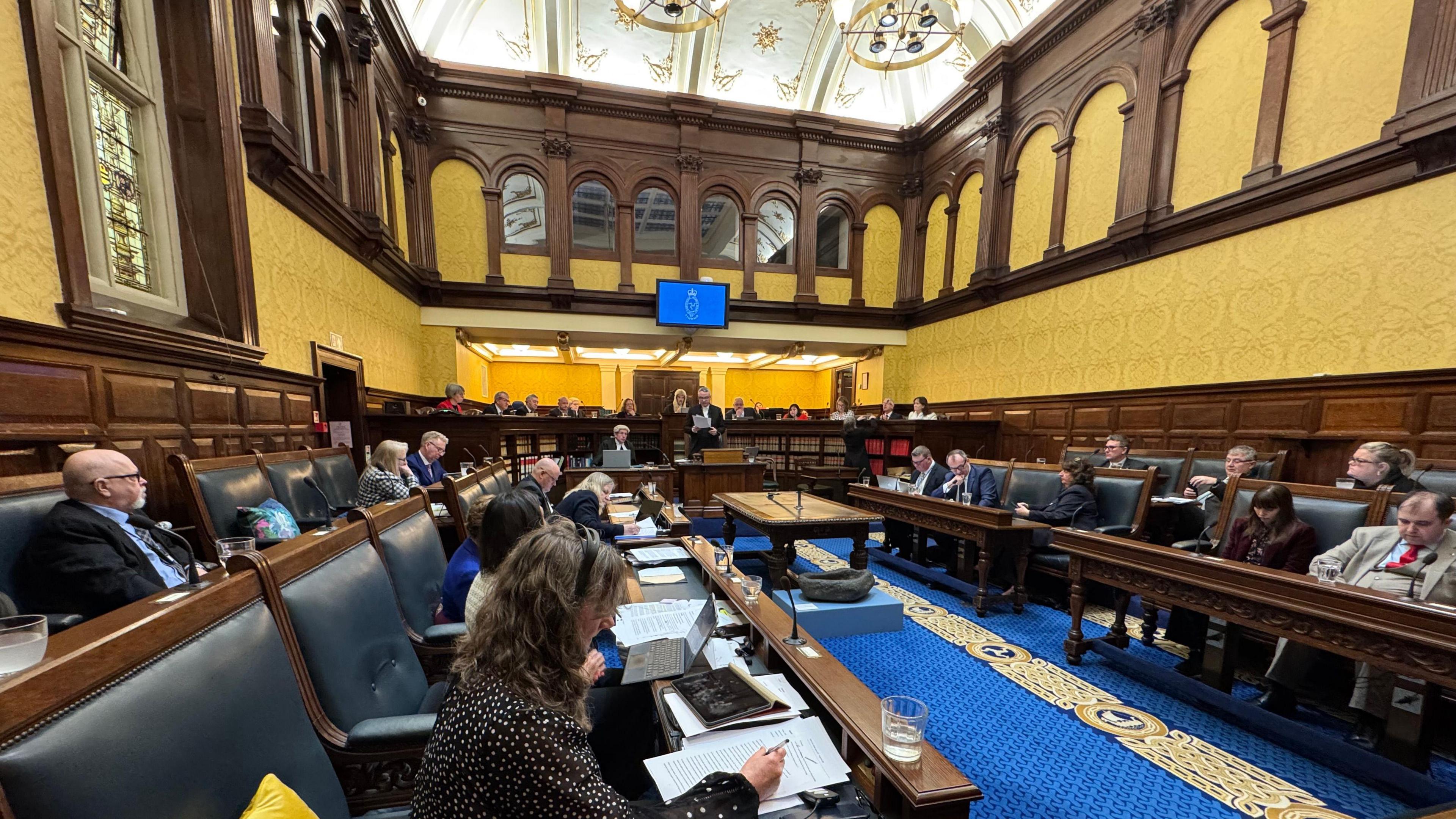 Members sitting in the Tynwald Chamber, which has wooden wall panels and benches with blue leather-clad seats and a blue carpet.