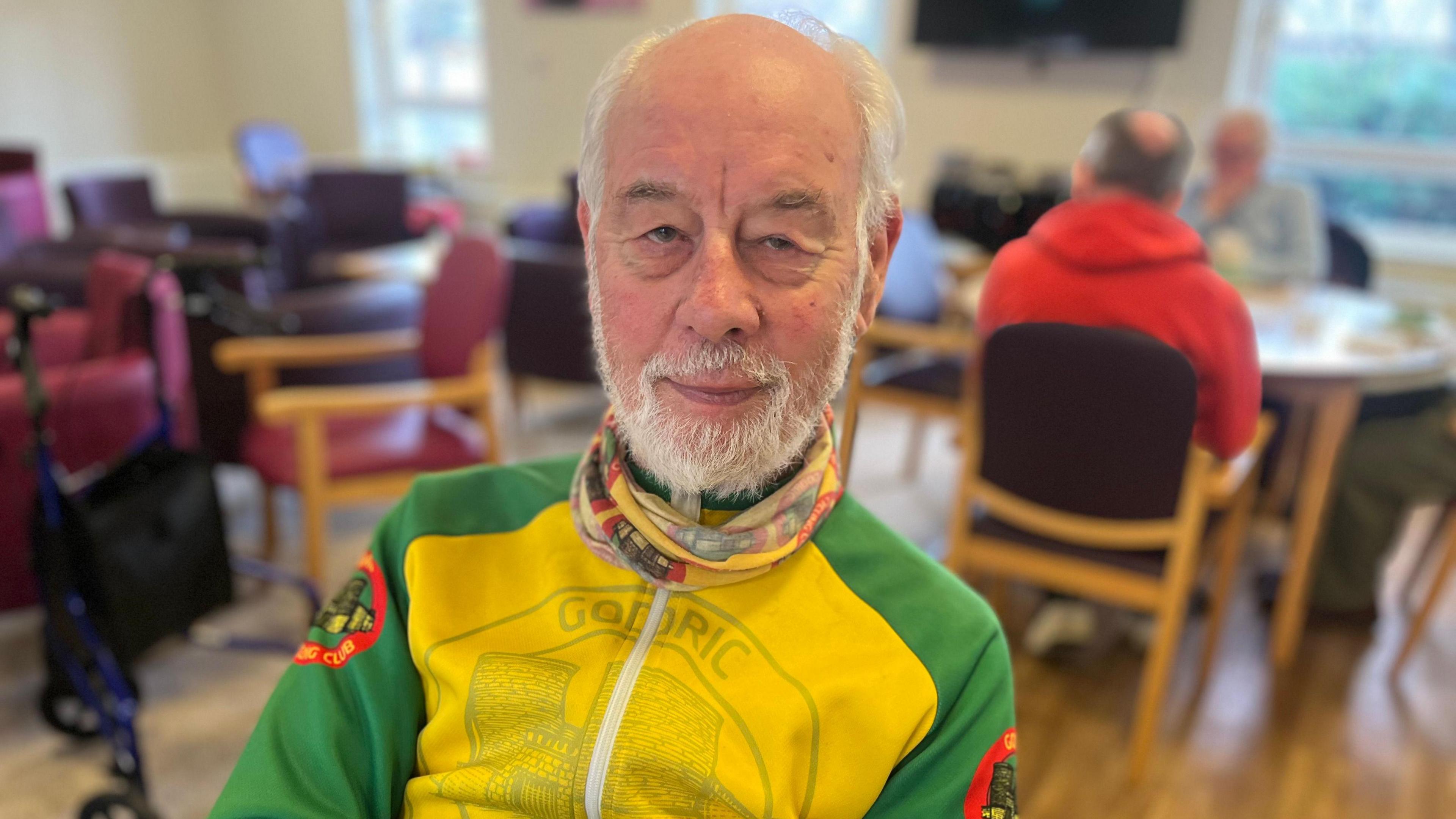 A cool older gentleman with a whispy white beard an a bald head smirks at the camera, wearing a green and yellow cycling top and a neck scarf.