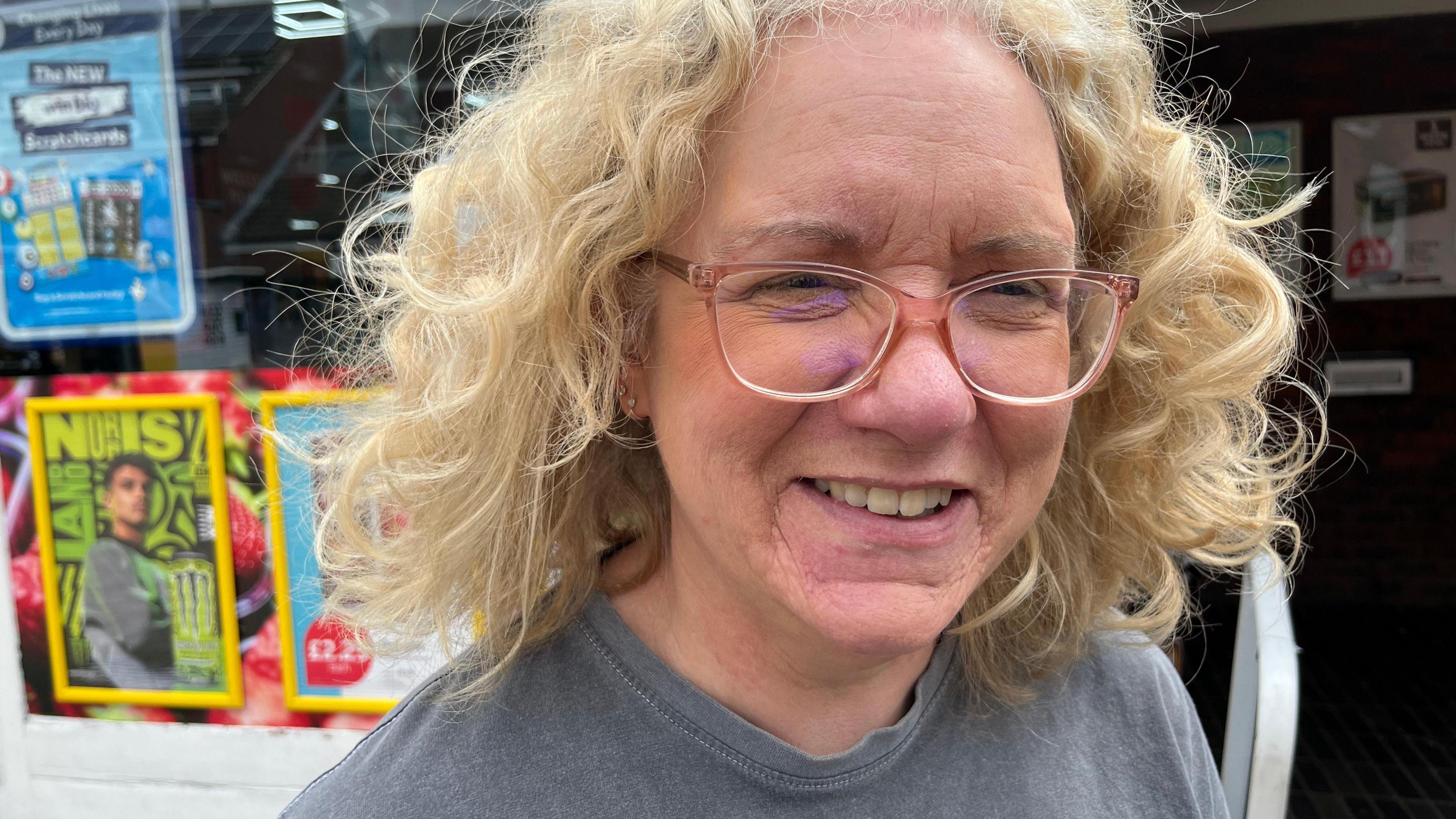 Tracey Westley is looking towards the camera and smiling. She wears light pink glasses and a grey t-shirt. Behind her is a window with advertisements stuck to it.