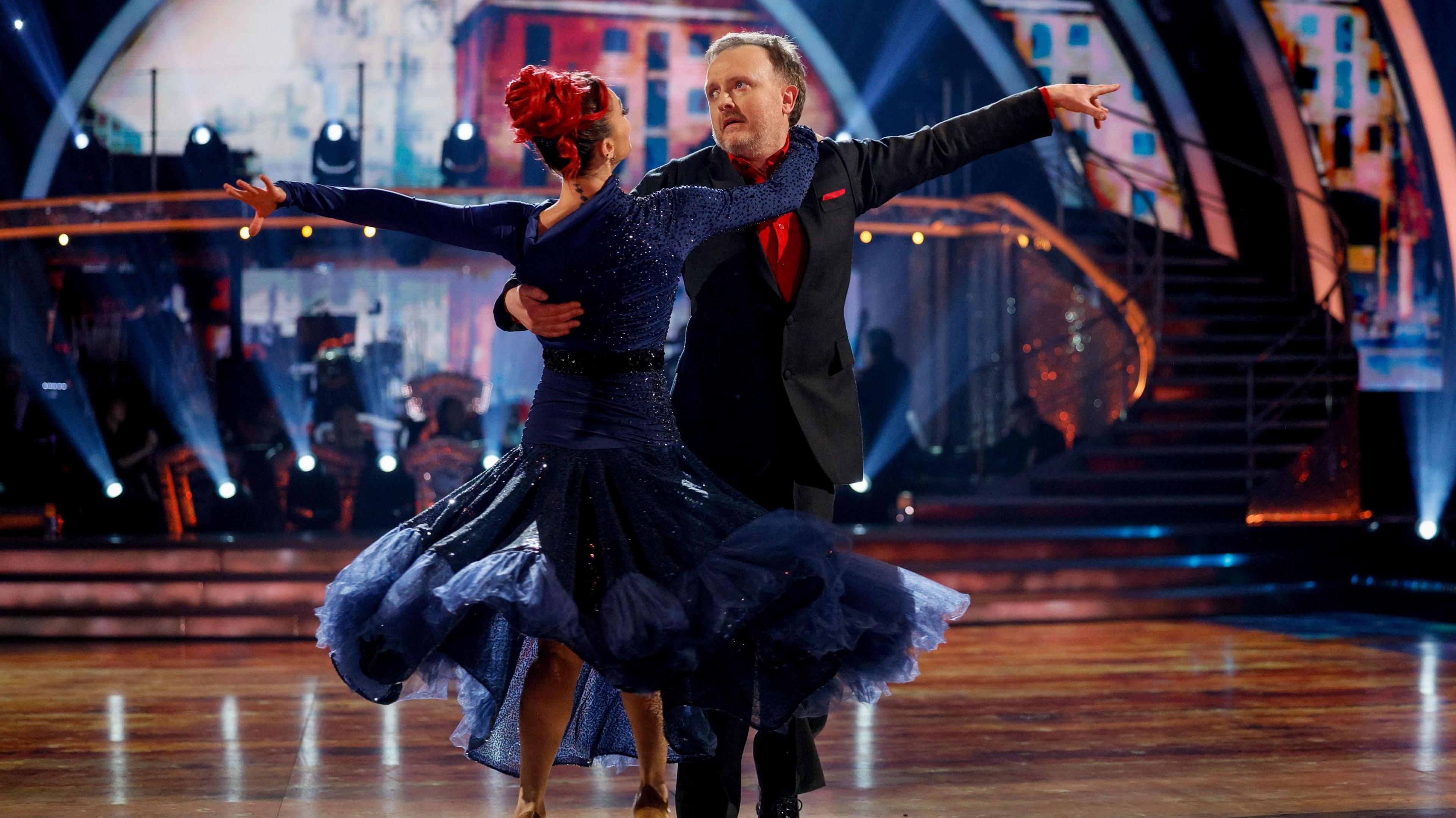 Chris McCausland, in a black blazer over a red shirt, dances with Diane Buswell. Both have their left arms outstretched behind them and hold on to each other with their right