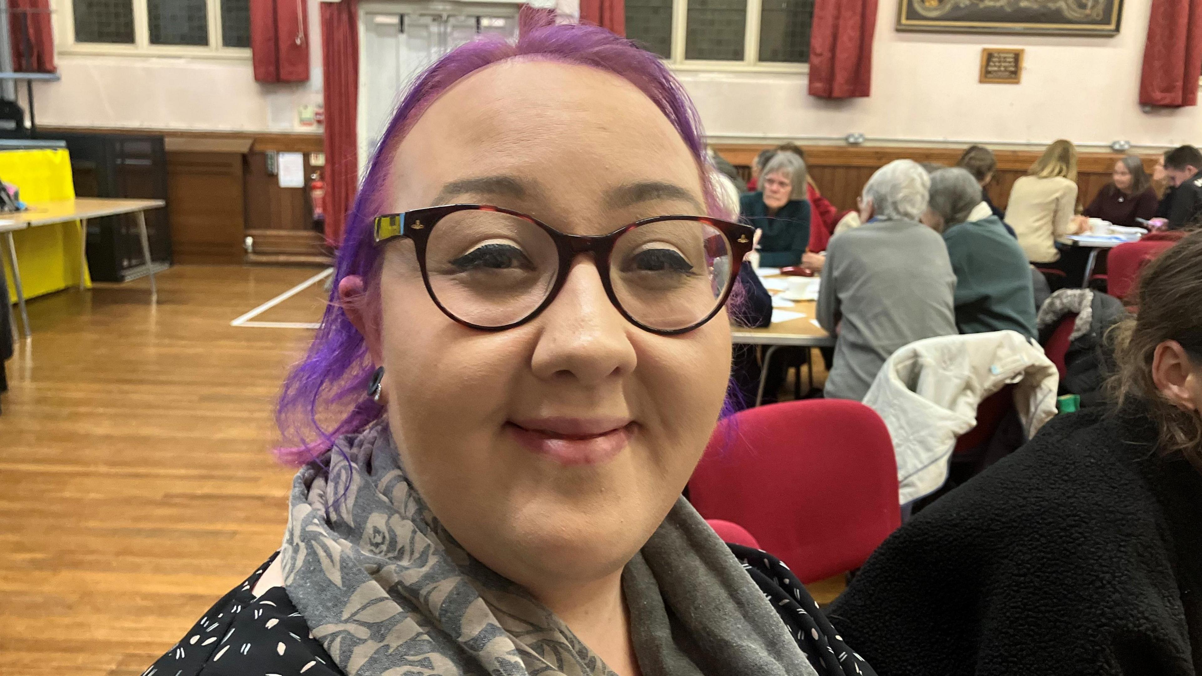 Woman with purple hair and glasses smiling at the camera. Behind her are people around tables discussing ideas 