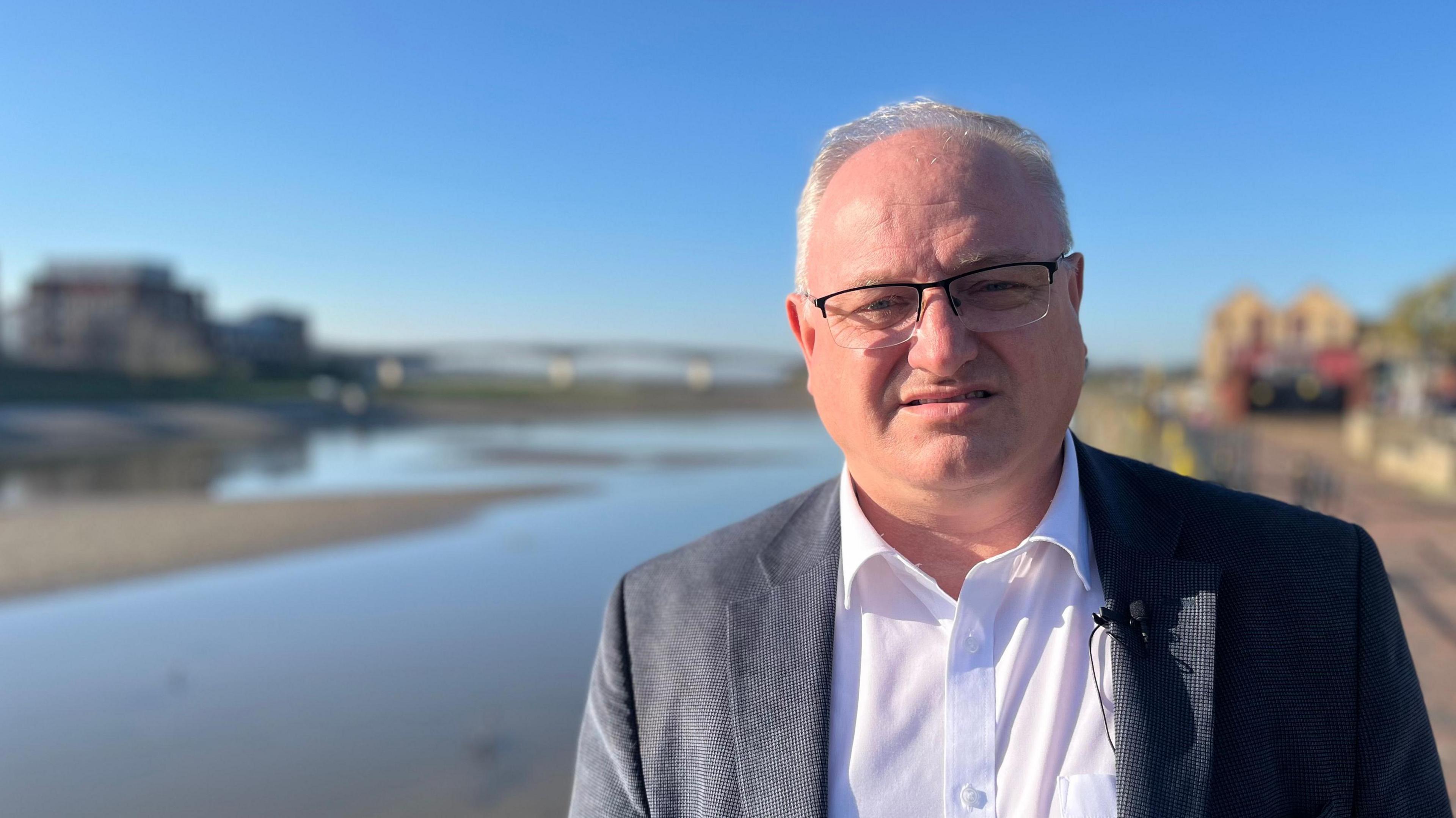 Ian Roome MP wearing a white shirt and a grey jacket, standing by the River Taw with the Taw Bridge in the background