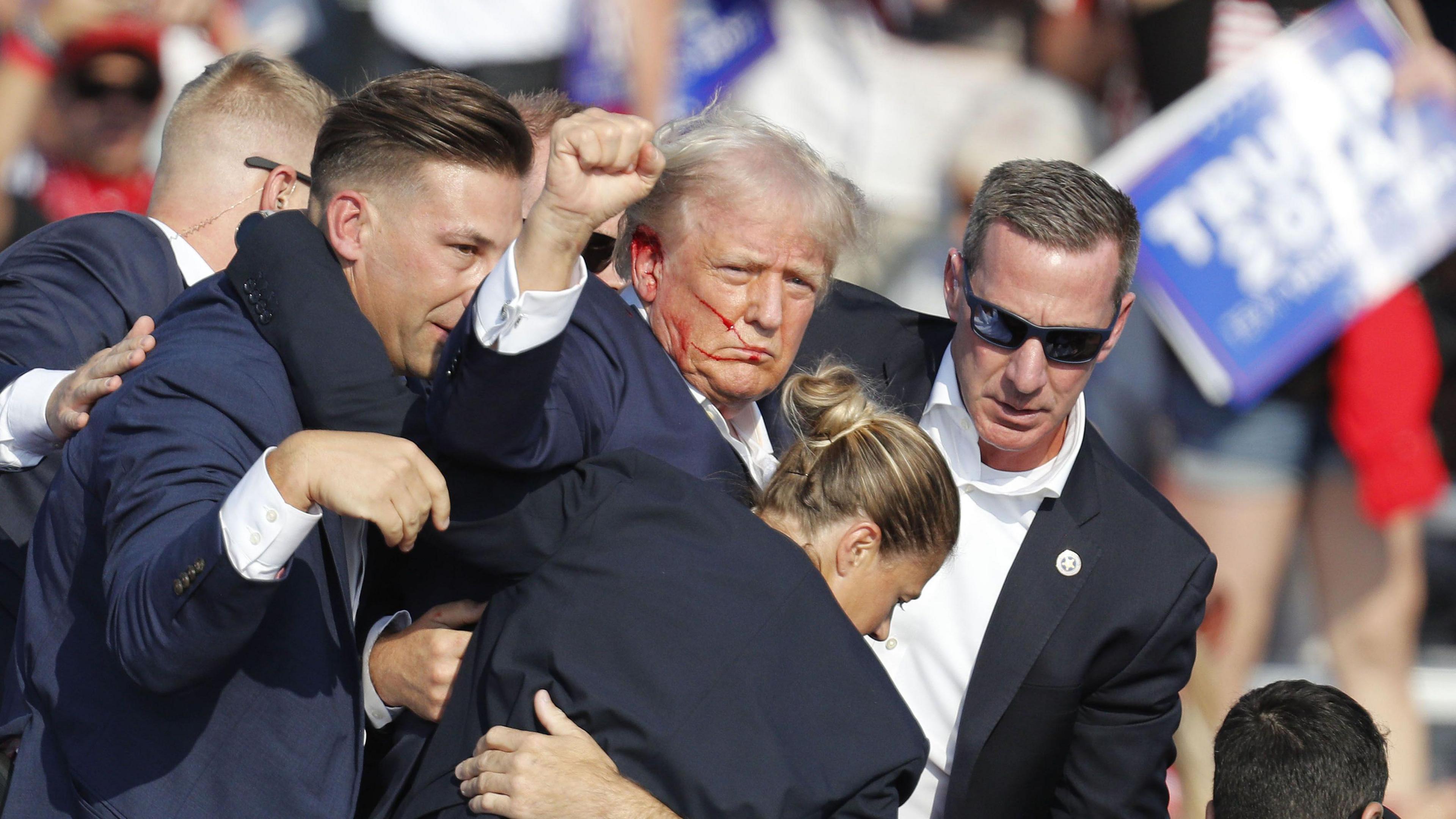 Donald Trump waving at fans with blood on his face, while being surrounded by his security team, who are all wearing dark suits.  