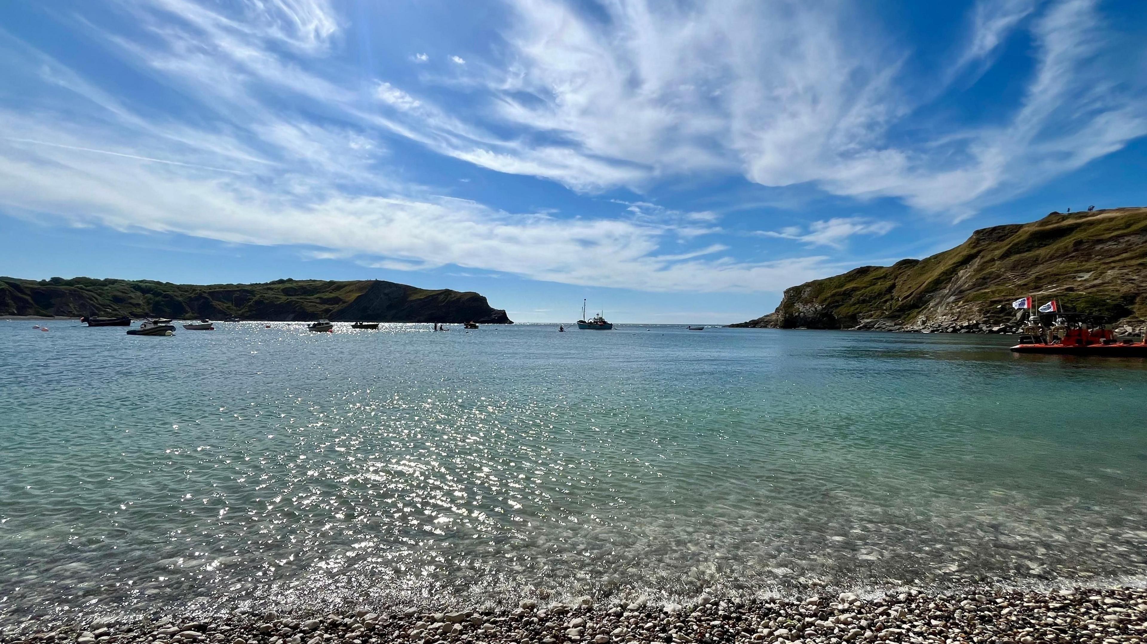 A cove at the seaside has several small boats moored. There are small ripples in the water on a sunny day under blue skies with some small white clouds. The cover is surrounded by low cliffs with green vegetation.