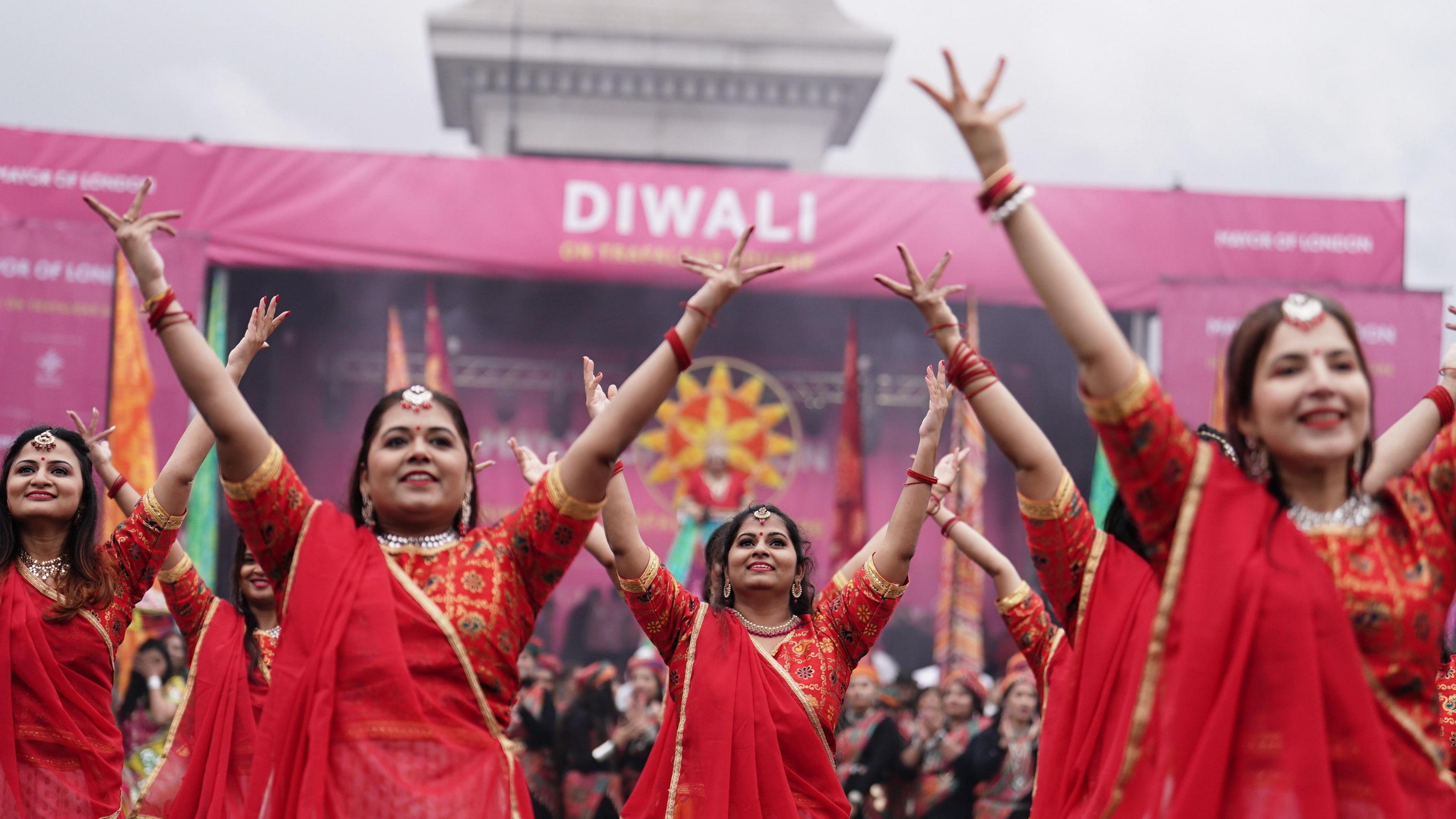 Four women standing in a dance formation and dressed in red and gold trimmed saris and wearing golden jewellery on their heads raise their hands in the air.  A stage behind them is decorated with purple banners: one says 'Diwali'. In the background is the plinth of Nelson's Column. 