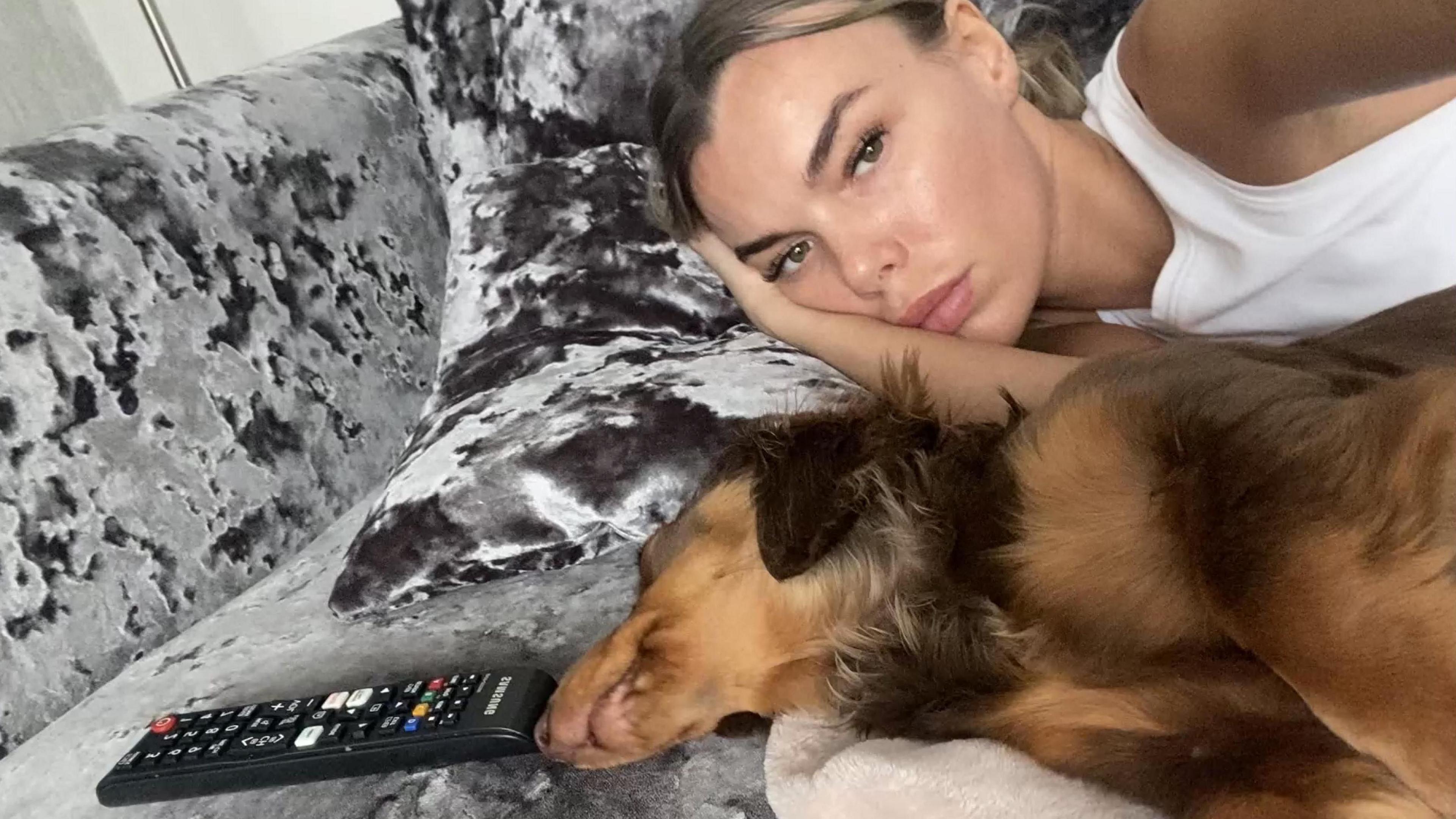 Ashley Dale, 28, with blonde hair and wearing a white vest top, lies on a grey couch with her brown long-haired Dachshund which is resting on its side.