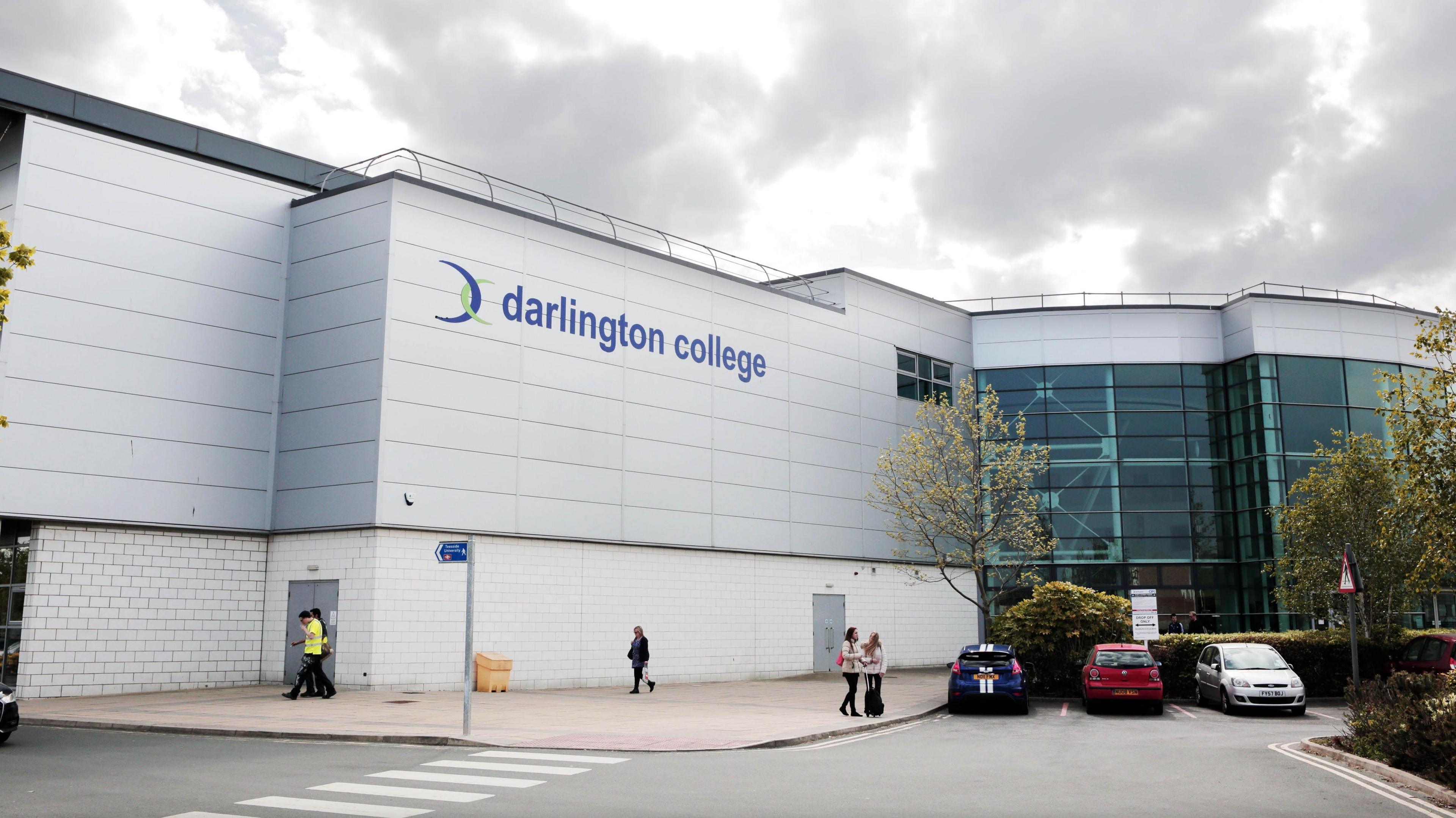 The outside of the Darlington College building, with the college's branding on the building. There is a grey sky above, and a few people walking around and some parked cars in the foreground. 