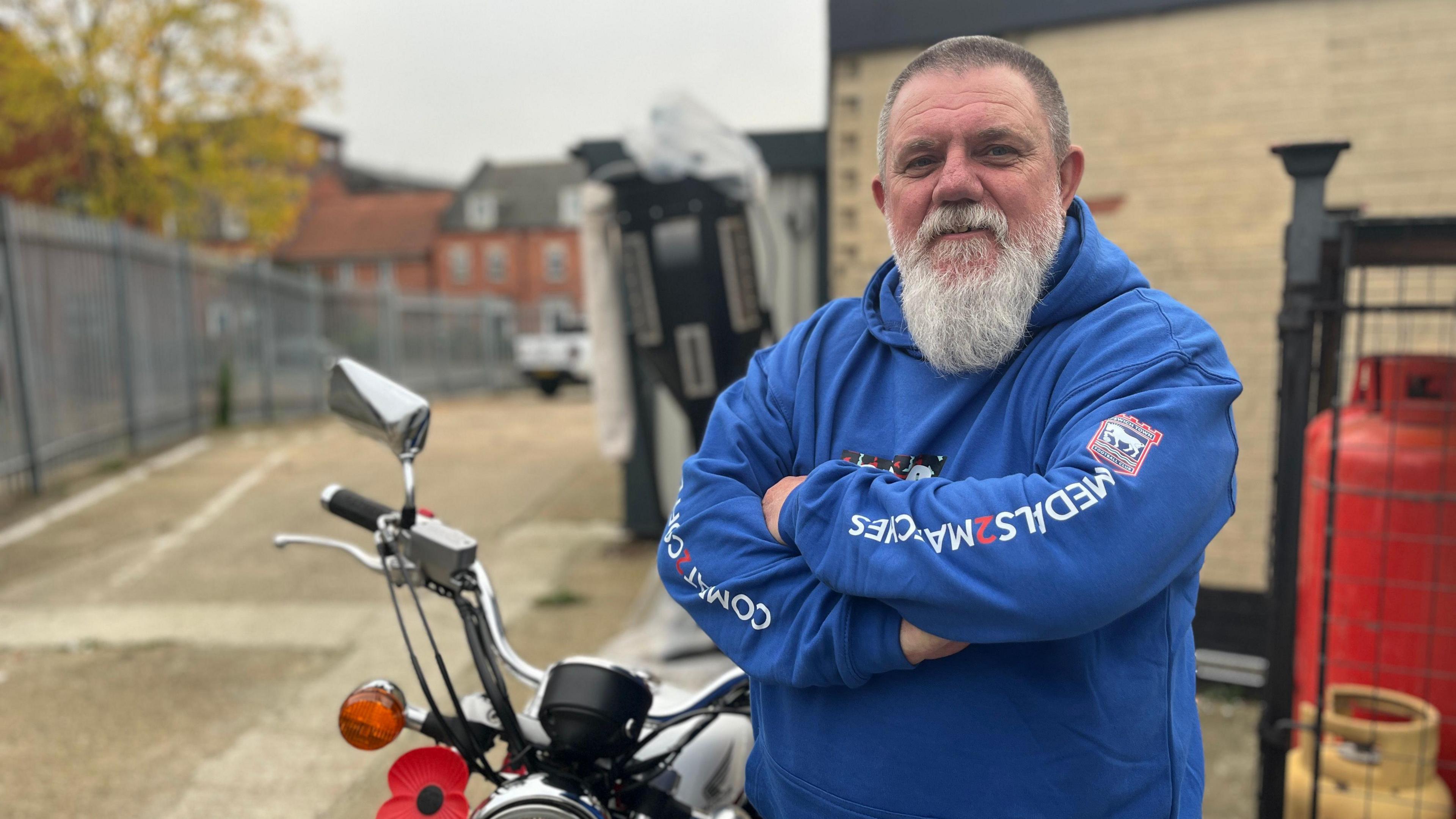 Paul Martin, wearing a blue hoodie, stands by an industrial unit next to a motorcycle which has a large poppy on the front. 