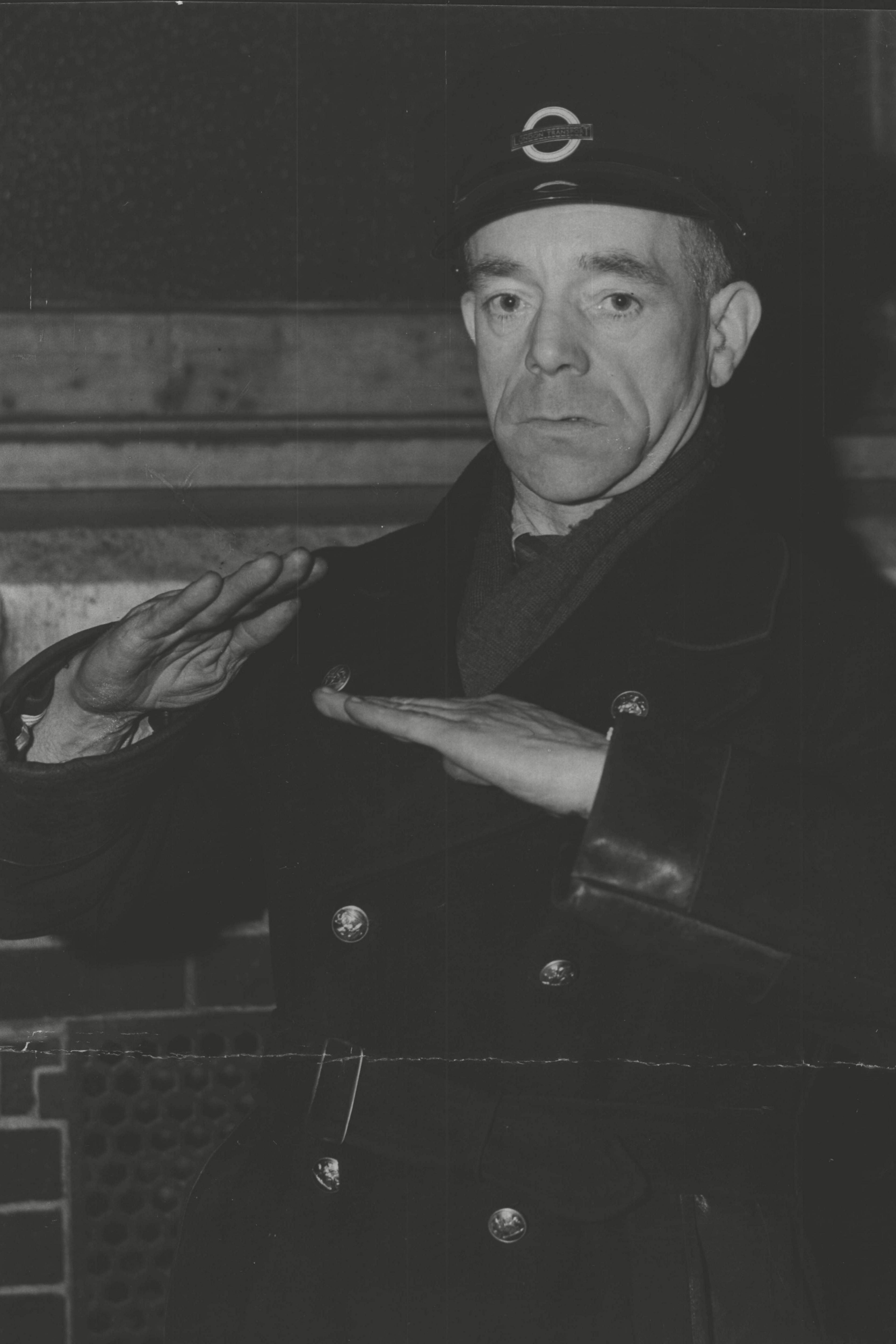 A black and white archive photo from 1952.  Albert Gunter is wearing his bus driver uniform, with a peaked cap with a badge on it, a double-breasted winter coat, and a muffler tucked in. He is unsmiling as he looks at the camera and has both hands at chest height, palms facing down. One hand is tilted above the other in a reconstruction of how Tower Bridge hinged open.