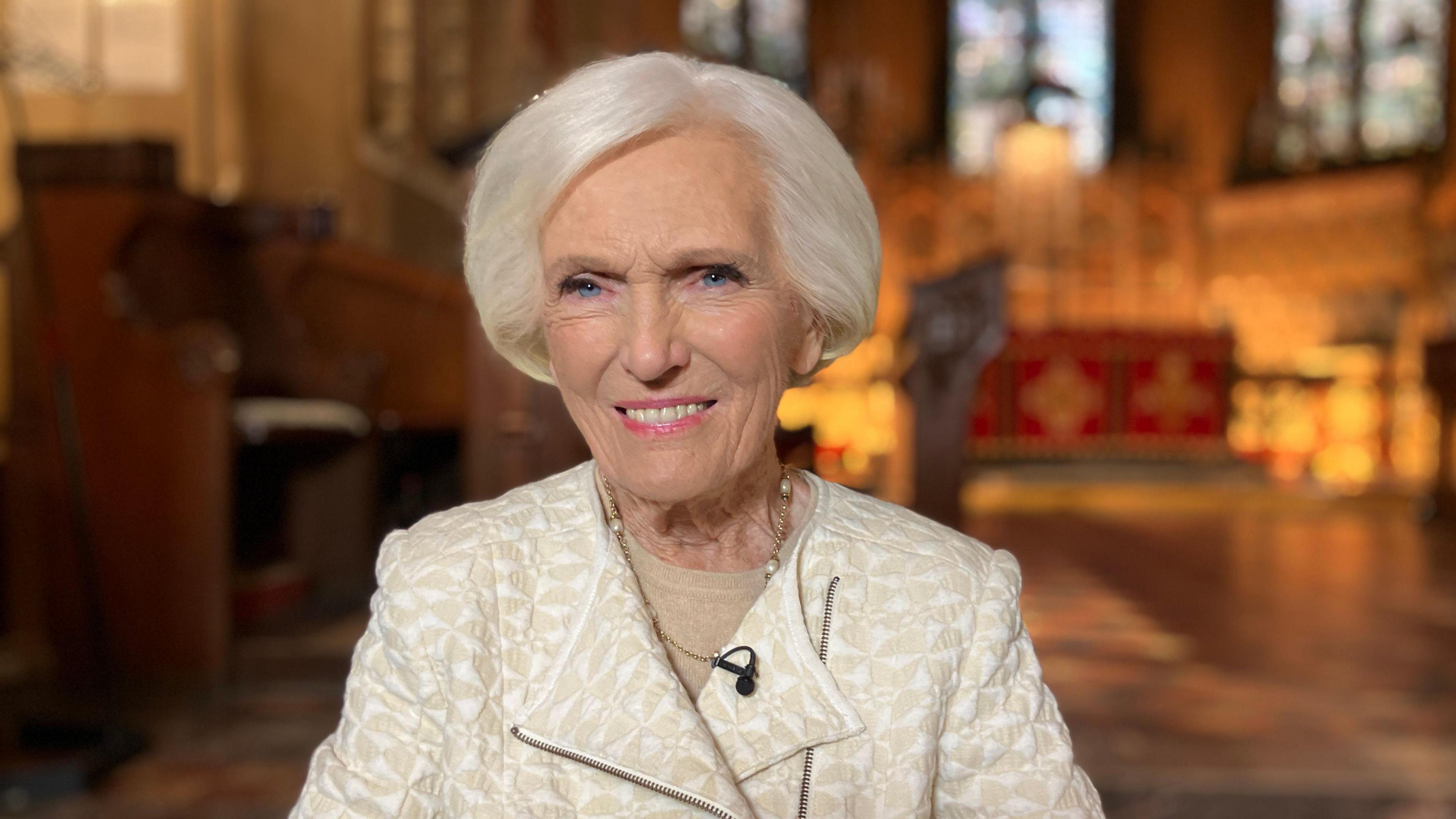 Mary Berry smiles in a large church, which is blurred in the background behind her. She is wearing a tailored white and cream jacket and short white hair. 