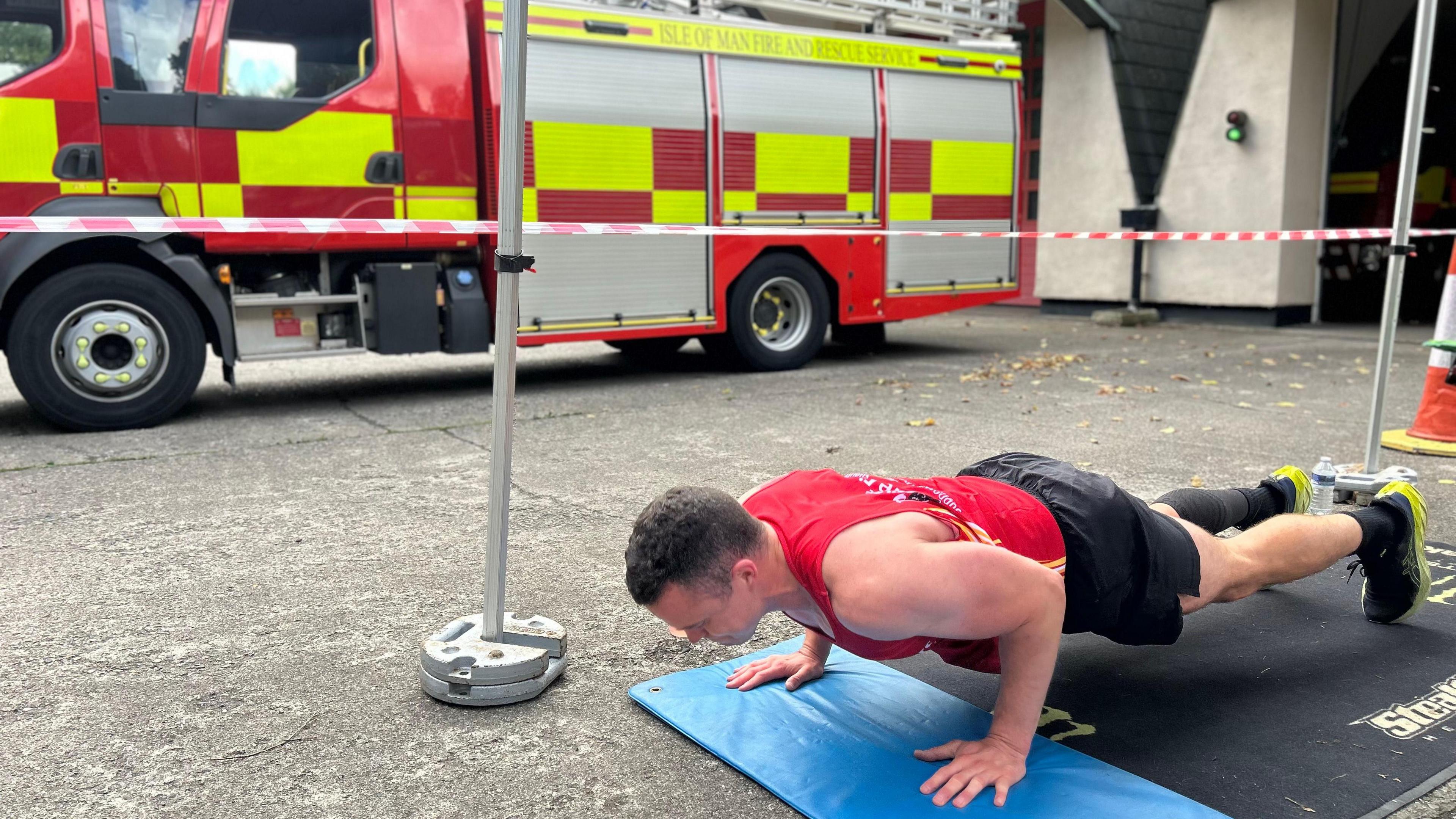 Adam Boyd wears a red vest, dark shorts and trainers. He is in a push up position on the forecourt of a fire station. A red and yellow fire engine can be seen in background.