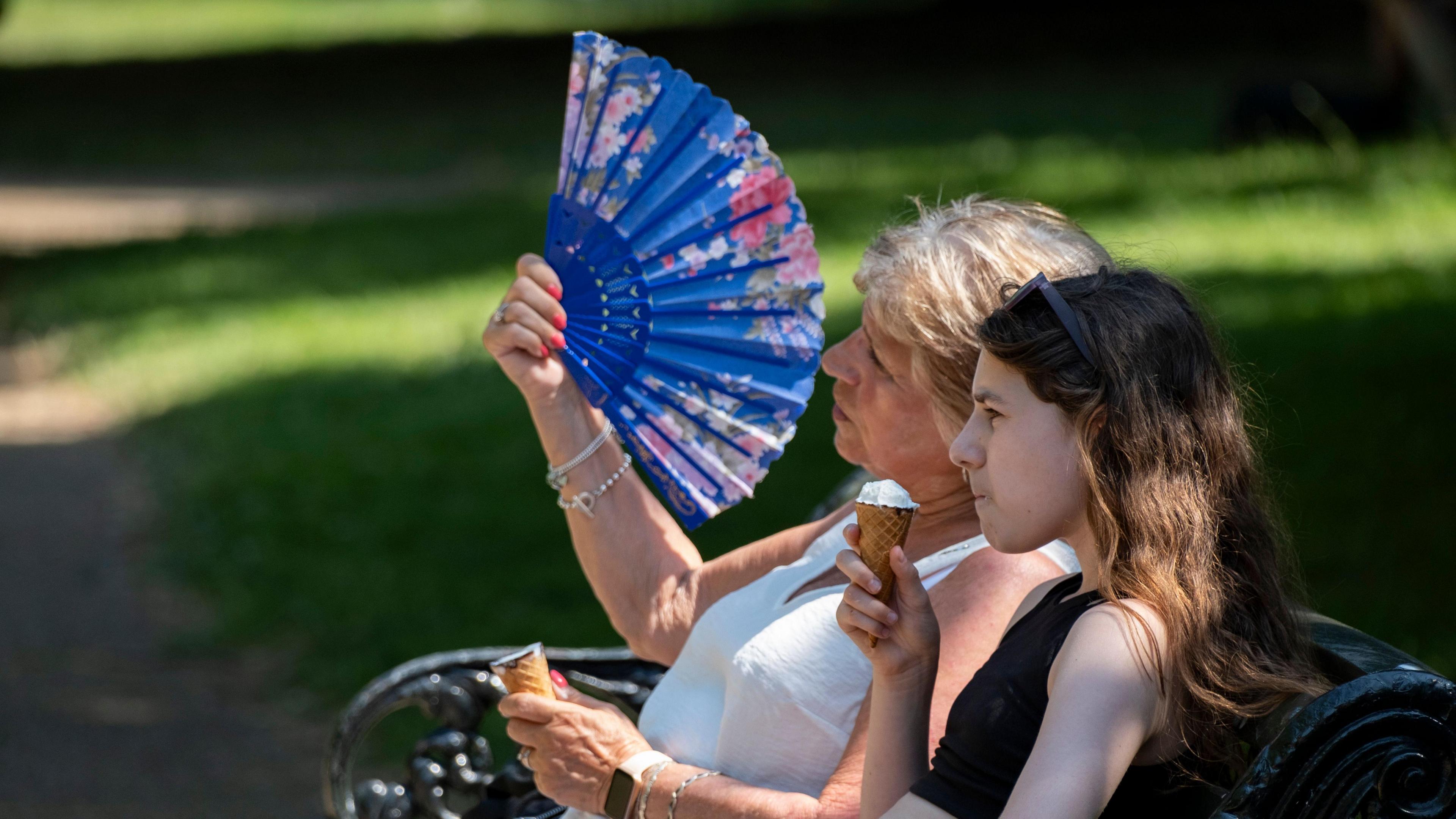 two women sitting on a park bench. One eating an ice cream, the other fanning herself in the hot weather