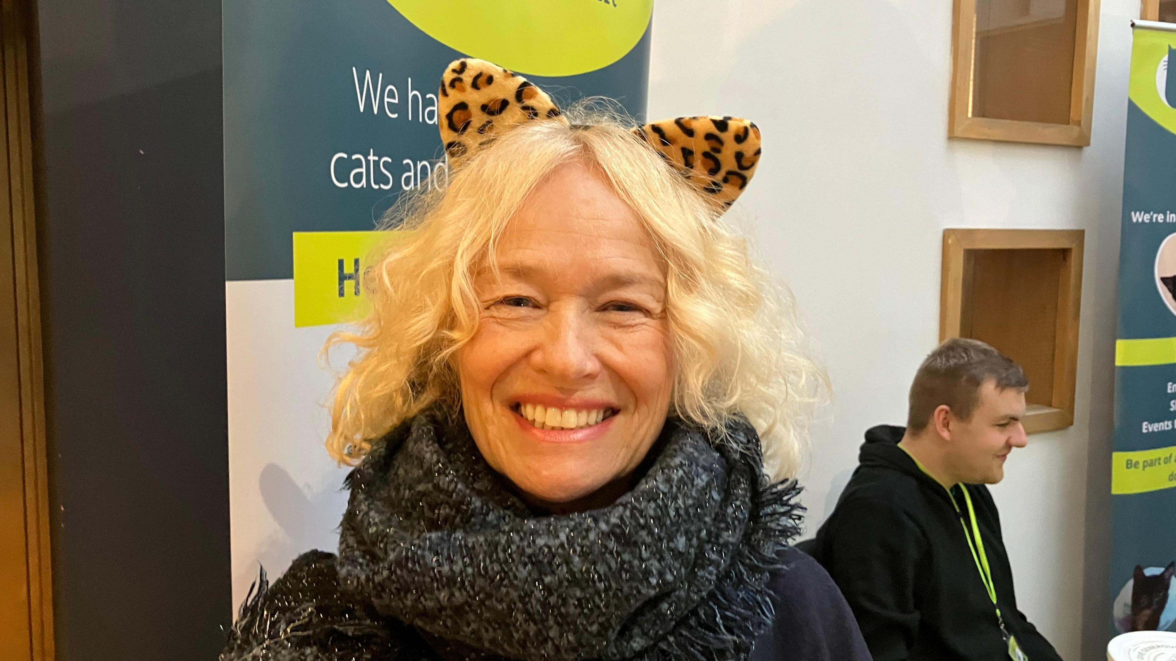 A woman with curly white hair wearing a pair of leopard print cat ears and a large glittery scarf.