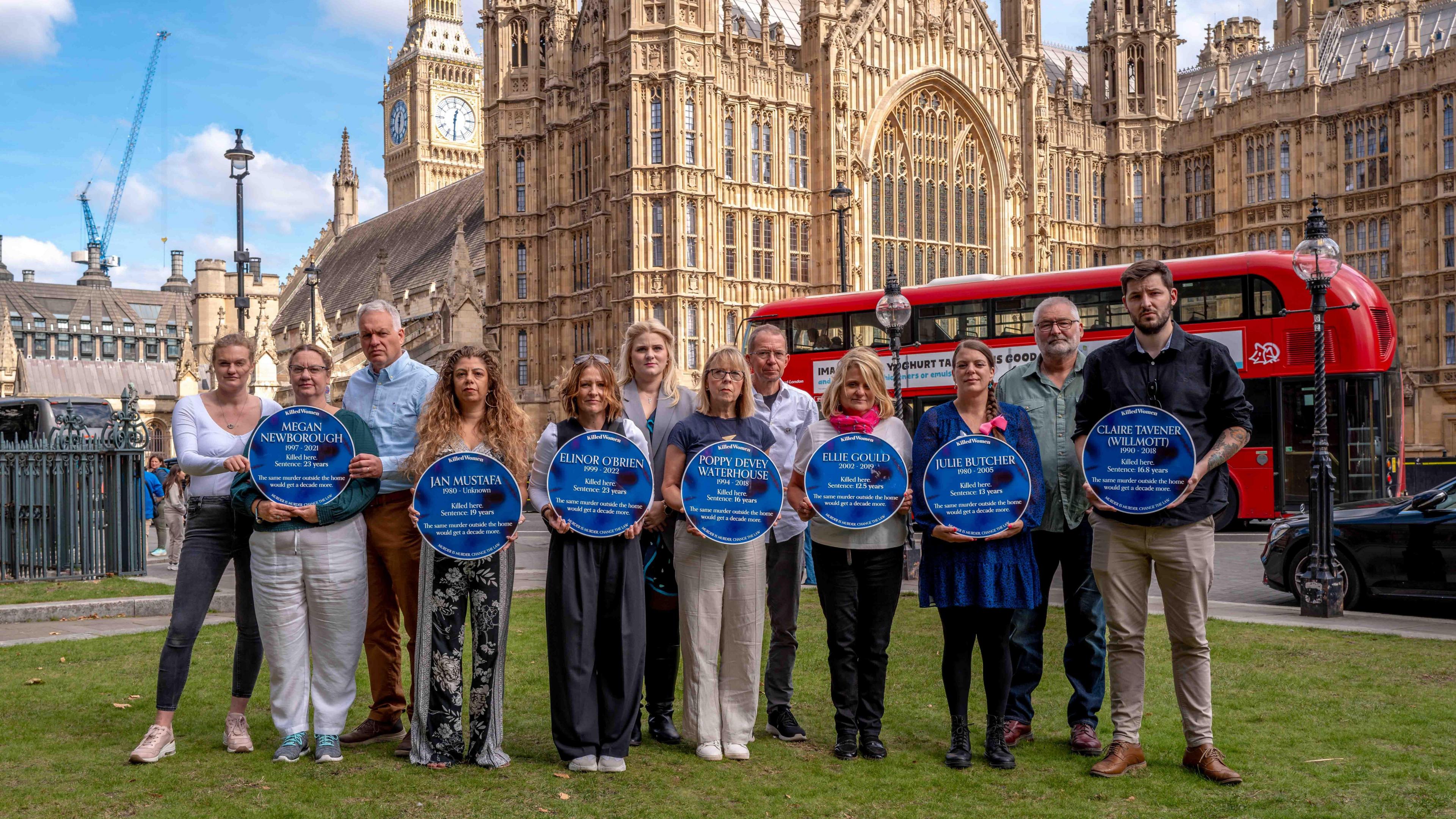 A group of people, some of whom are holding blue discs which are peppered with black marks, stand on a patch of grass in front of the Houses of Parliament as a red bus goes past.