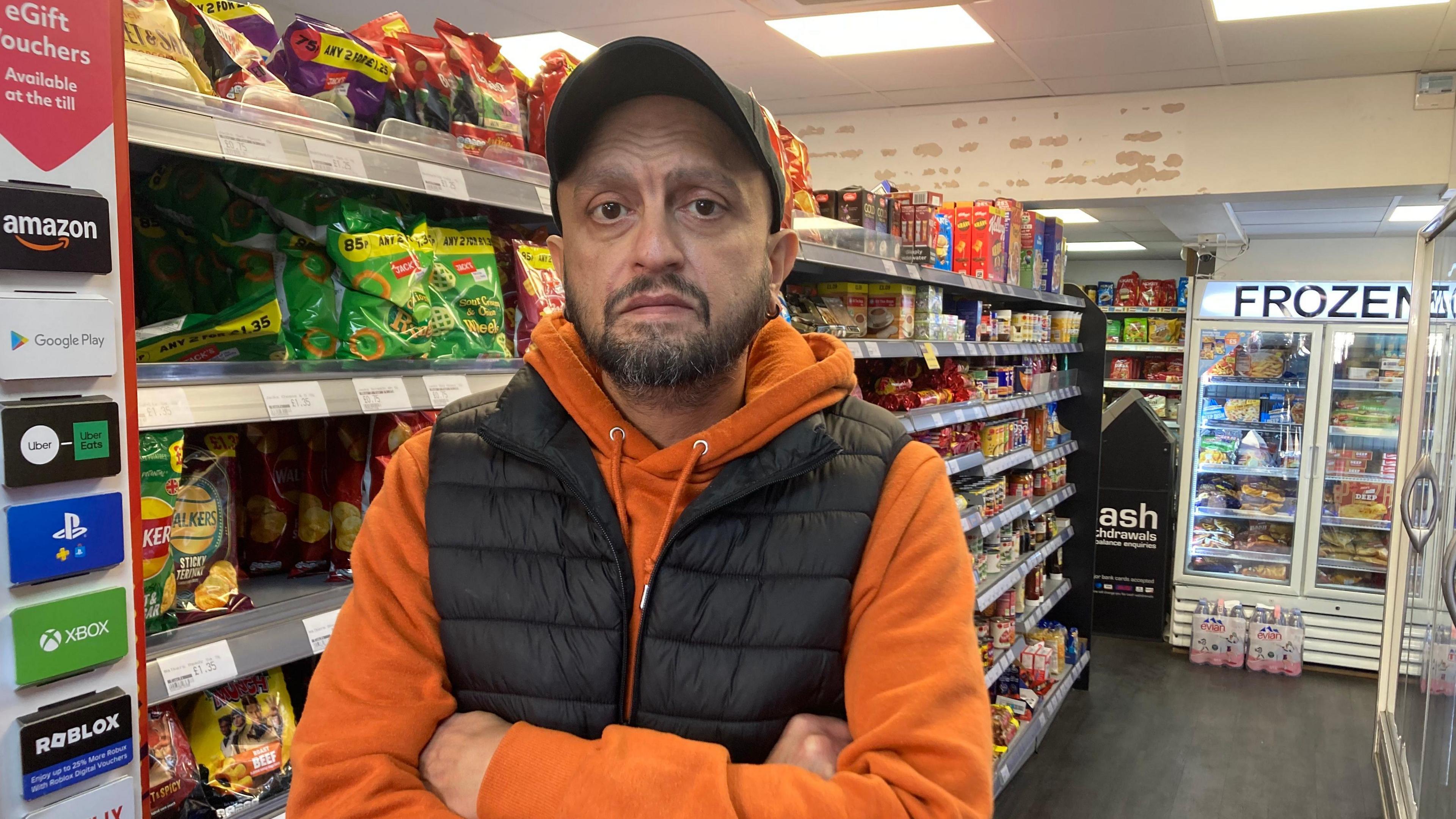 A man stands in the aisle of a convenience store with shelves full of crisps and other products behind him. He has brown eyes and a short brown beard. He is frowning with his arms crossed and wearing an orange hoodie, black gillet and black cap.